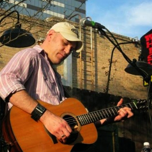 Person playing guitar outdoors, wearing a cap and striped shirt, with microphone and brick wall background.