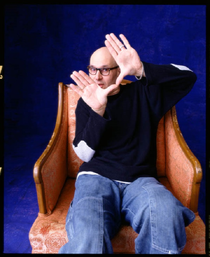 Person with glasses sitting in a chair, hands raised, against a dark blue background.