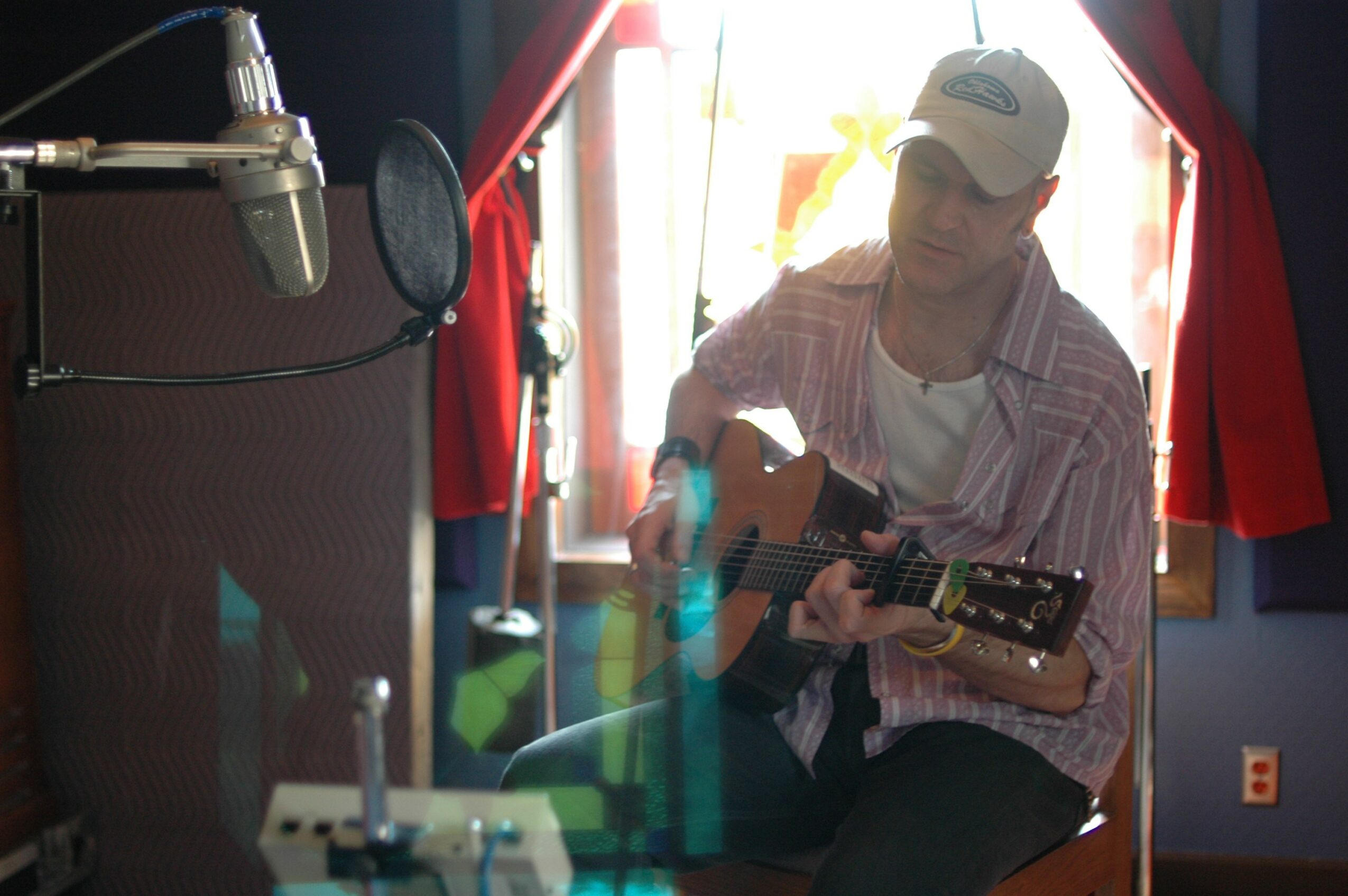 Person wearing a cap playing an acoustic guitar in a room with red curtains and a window behind.