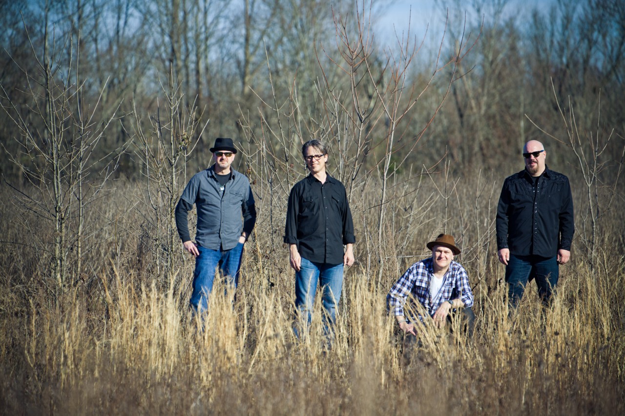 Four people standing and sitting in a grassy field with trees in the background, sunny day.