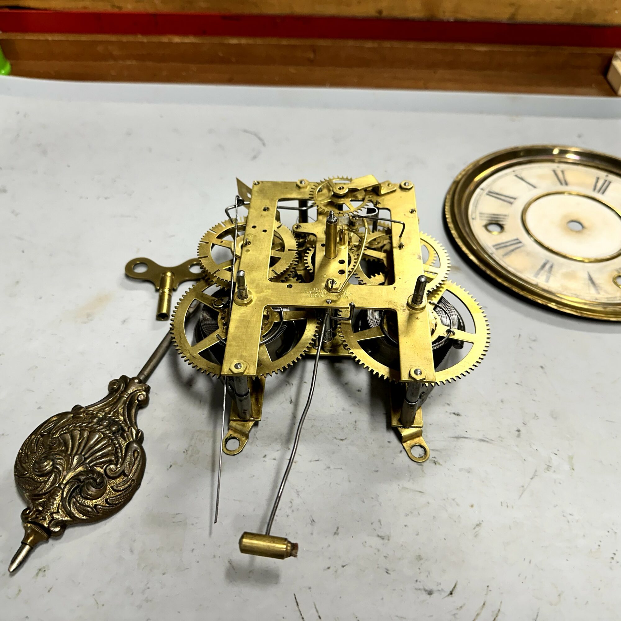 Disassembled clock mechanism with gears, pendulum, and decorative clock face on a white surface.