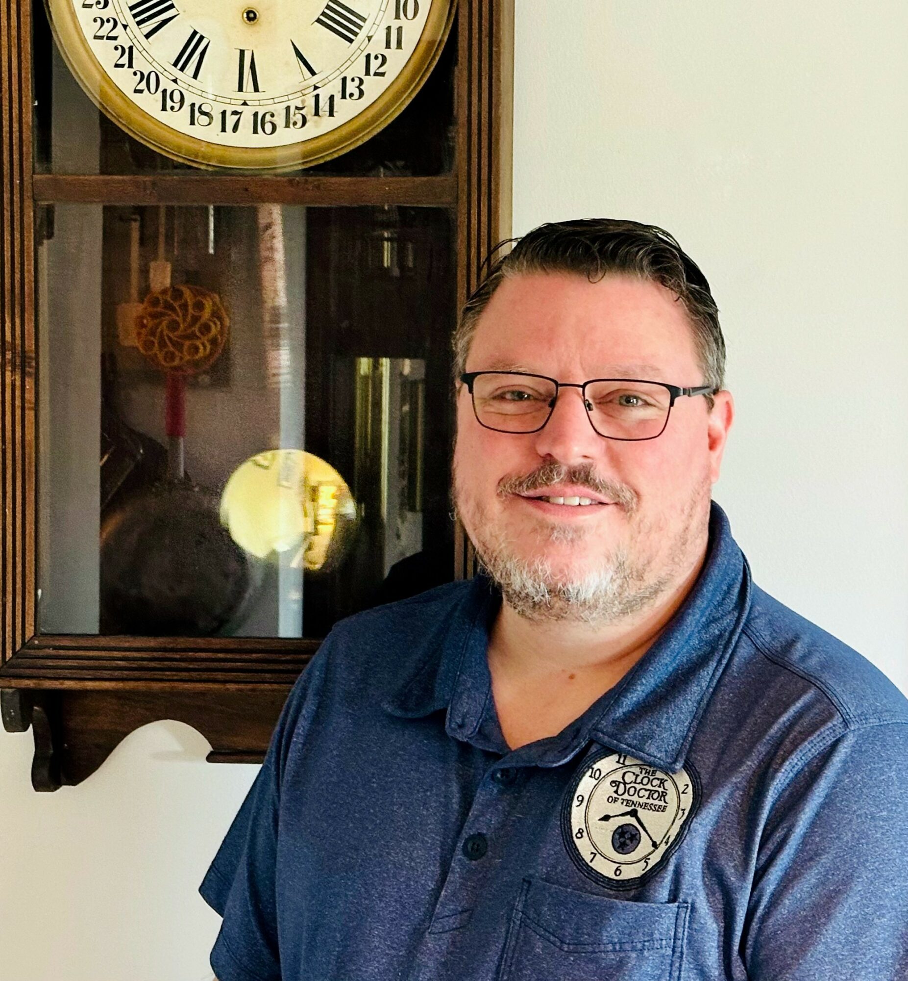 Man with glasses and short hair, wearing a blue shirt with a badge, smiling indoors near a clock and cabinet.