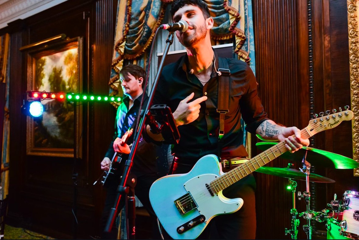 Two musicians perform on stage with guitars, microphone, and colorful lighting, in a wood-paneled room with paintings and curtains.