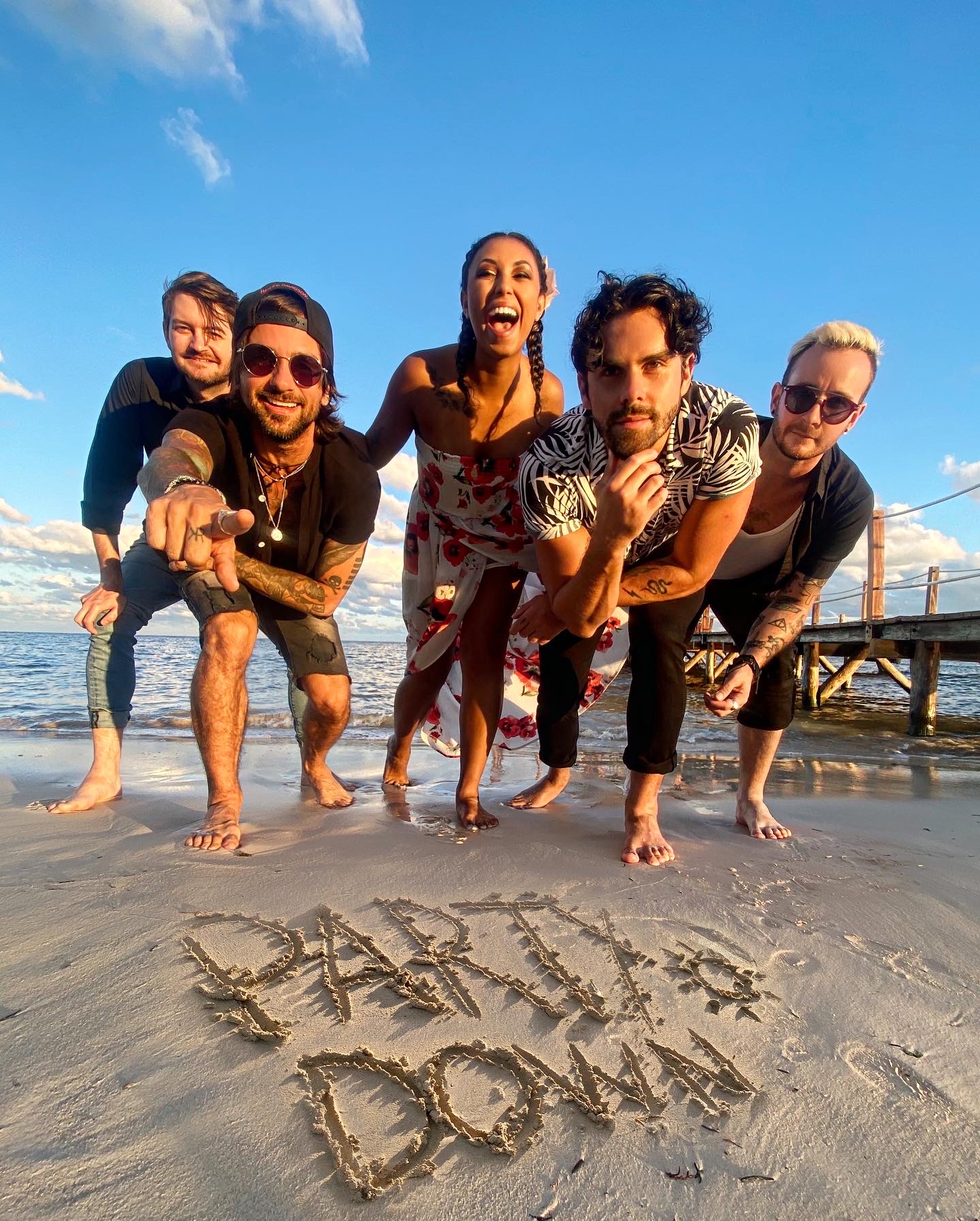 Group of five people on a beach, smiling and posing behind the words 'BABY DOLL' written in the sand.