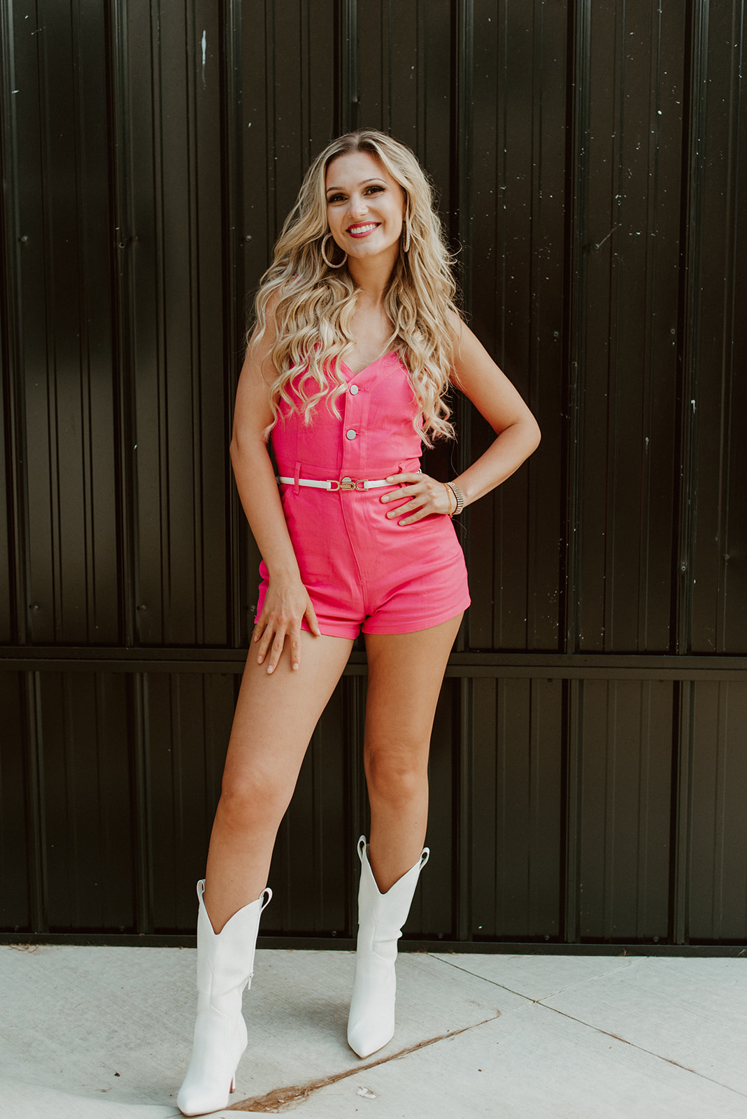 Young woman with long wavy blonde hair in pink romper and white cowboy boots, standing outdoors in front of a dark wooden fence.