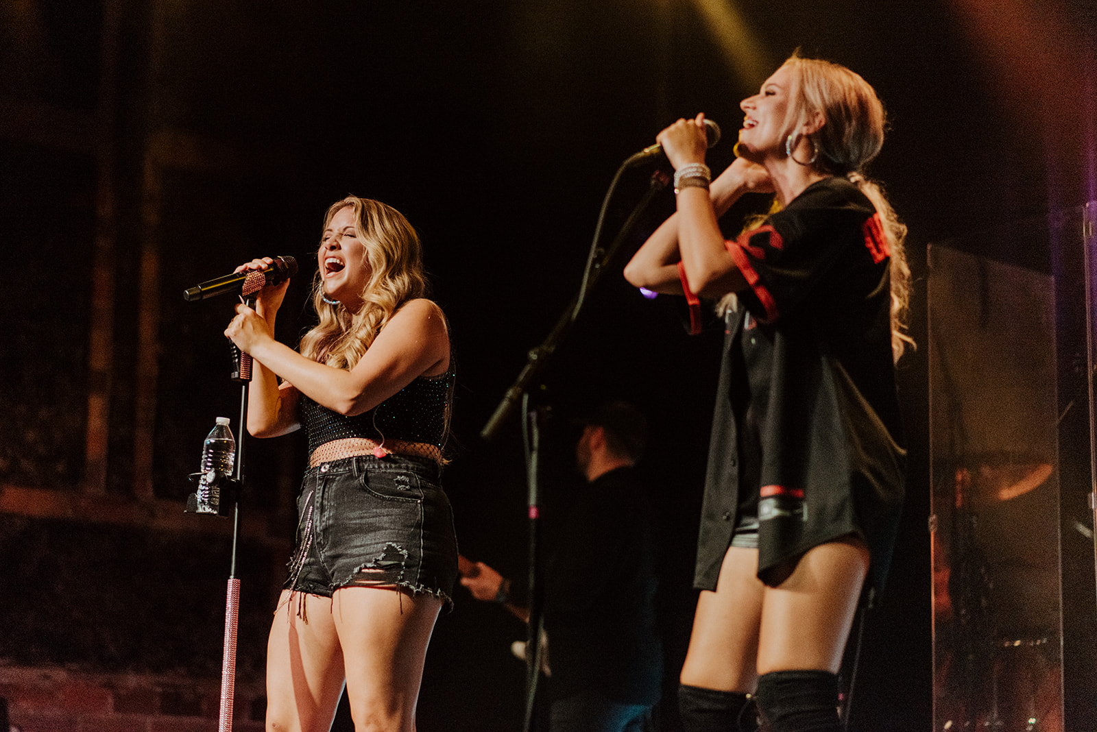 Two women singing on stage with microphones, one with long hair and the other with short hair, in a concert setting.