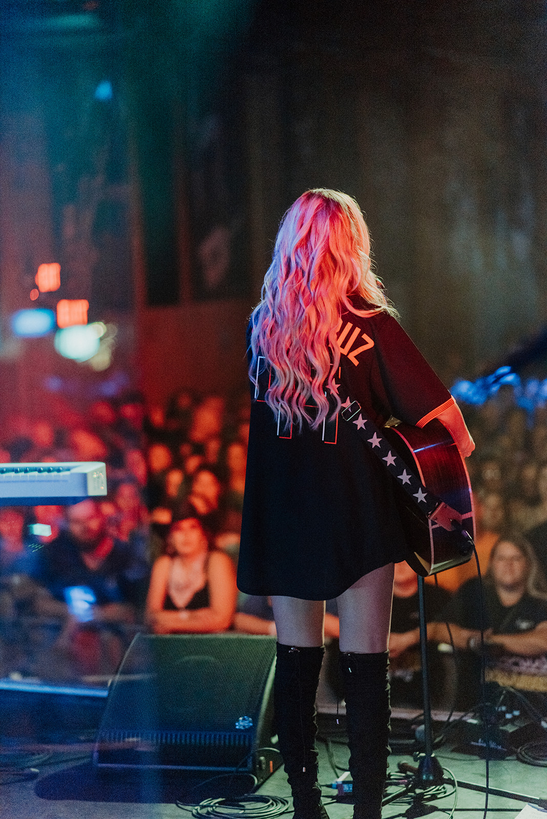 Performer with long, wavy pink hair on stage facing an audience, holding a guitar, with colorful stage lighting.