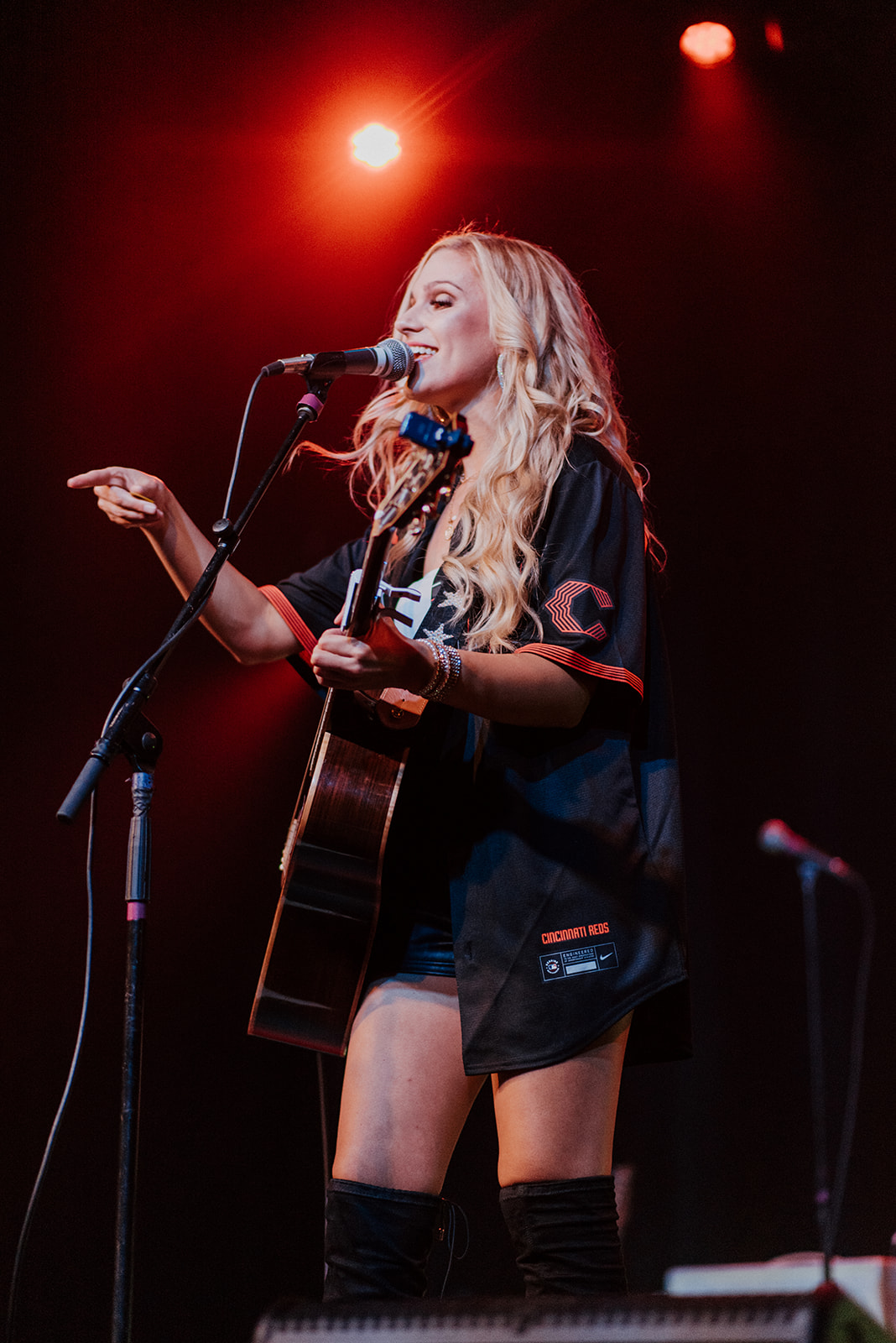 Young woman with long blonde hair singing into a microphone while playing an acoustic guitar on stage.
