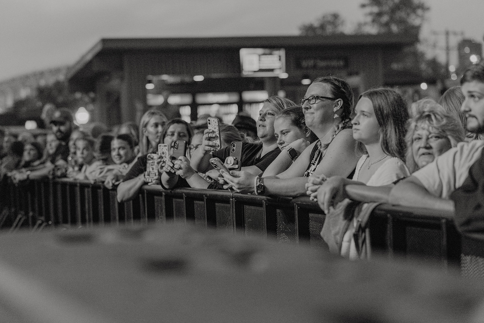 Crowd of people standing behind a barrier, watching an event outdoors, some taking photos or videos.