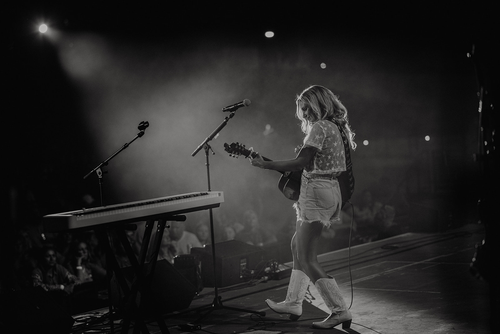 Person playing guitar on stage with microphone and keyboard nearby, audience in background, stage lighting illuminating scene.