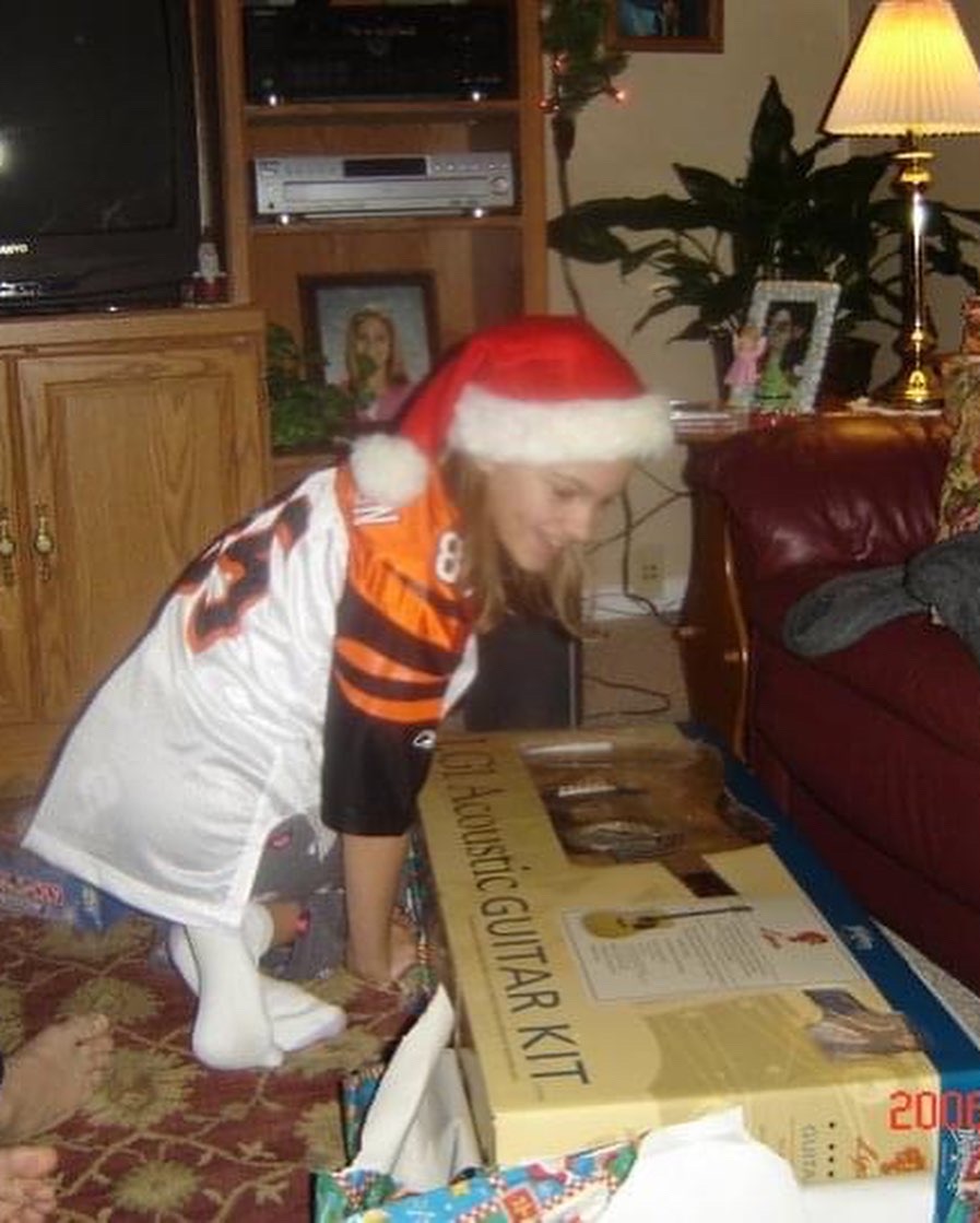 Child wearing a Santa hat and sports jersey, bending over a large guitar kit box in a living room.