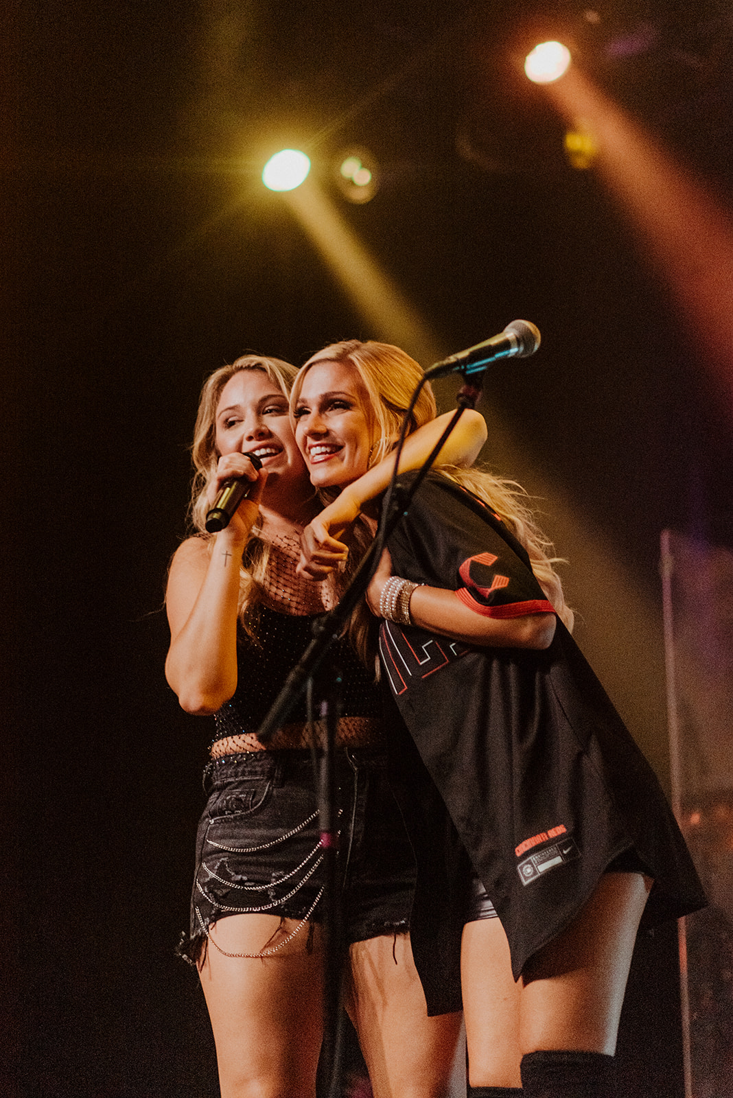 Two women smiling and hugging on stage with microphones, stage lights shining above them.
