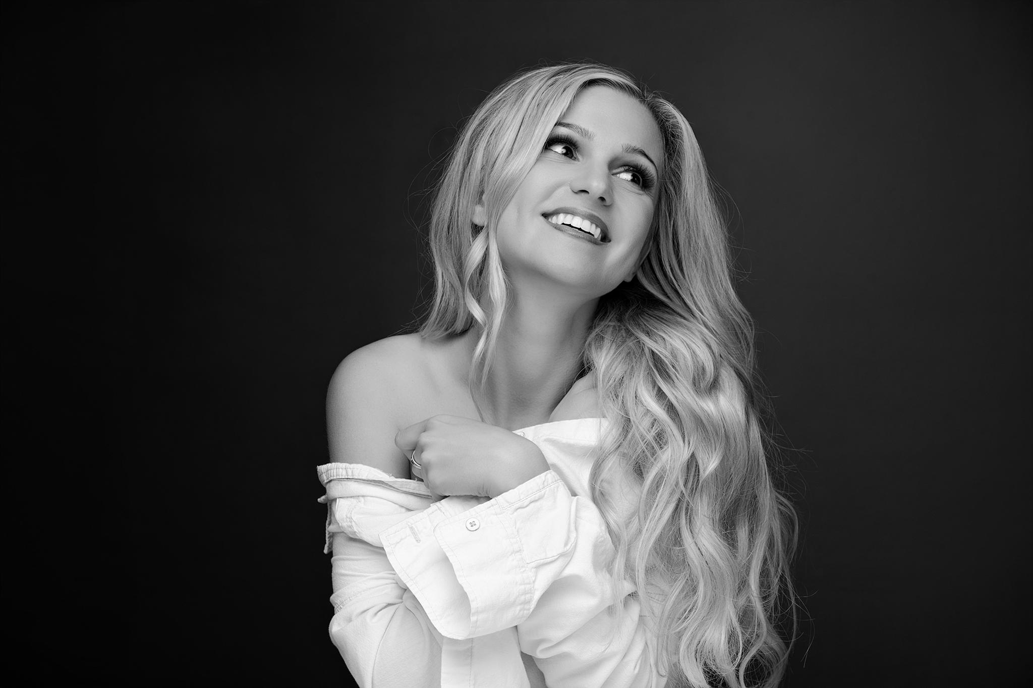 Woman with long wavy hair smiling and looking upward, wearing an off-shoulder top, black background.
