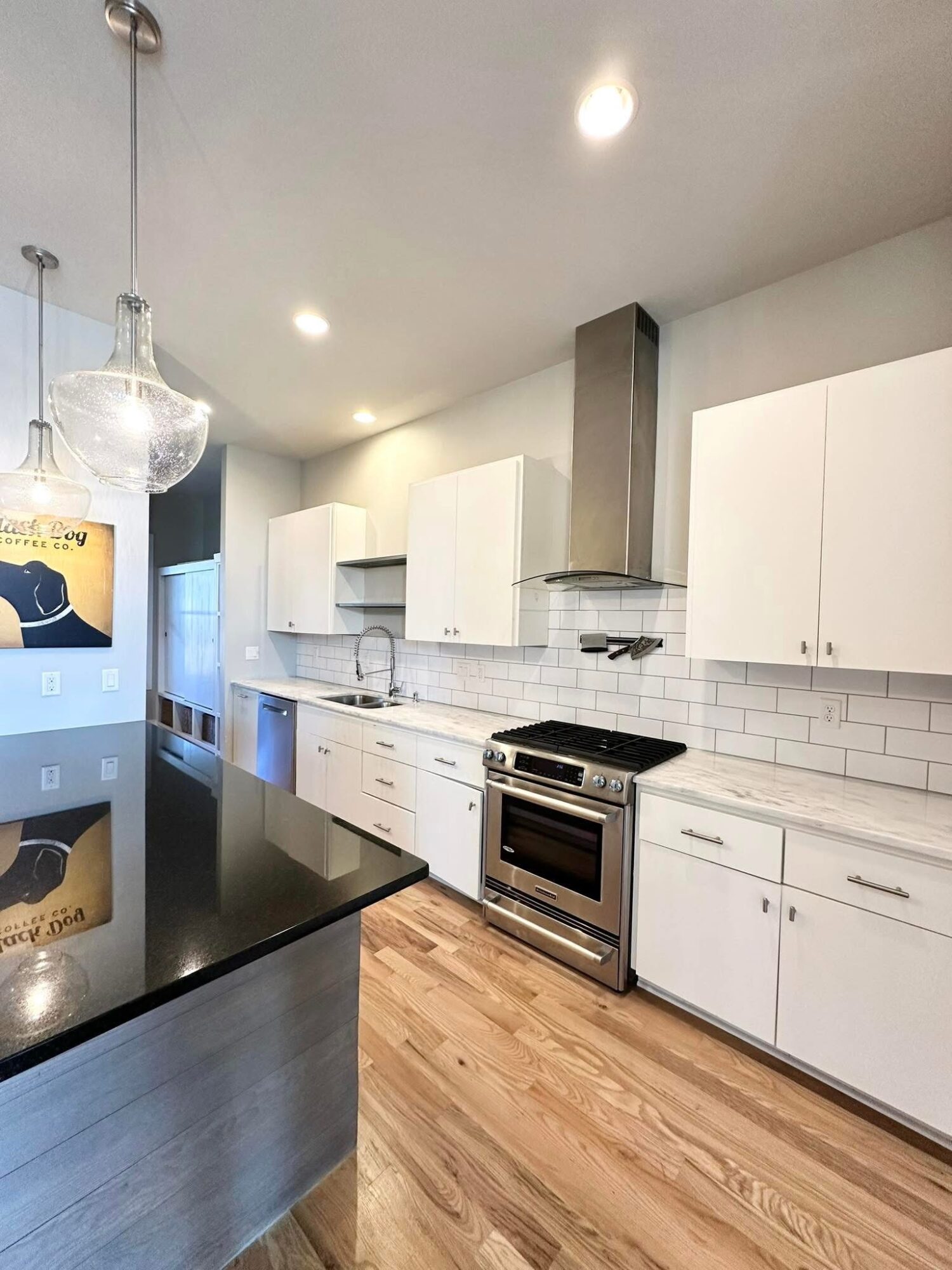 Modern kitchen with white cabinets, stainless steel oven, black countertop, and wooden floor, illuminated by ceiling lights and pendant lamps.