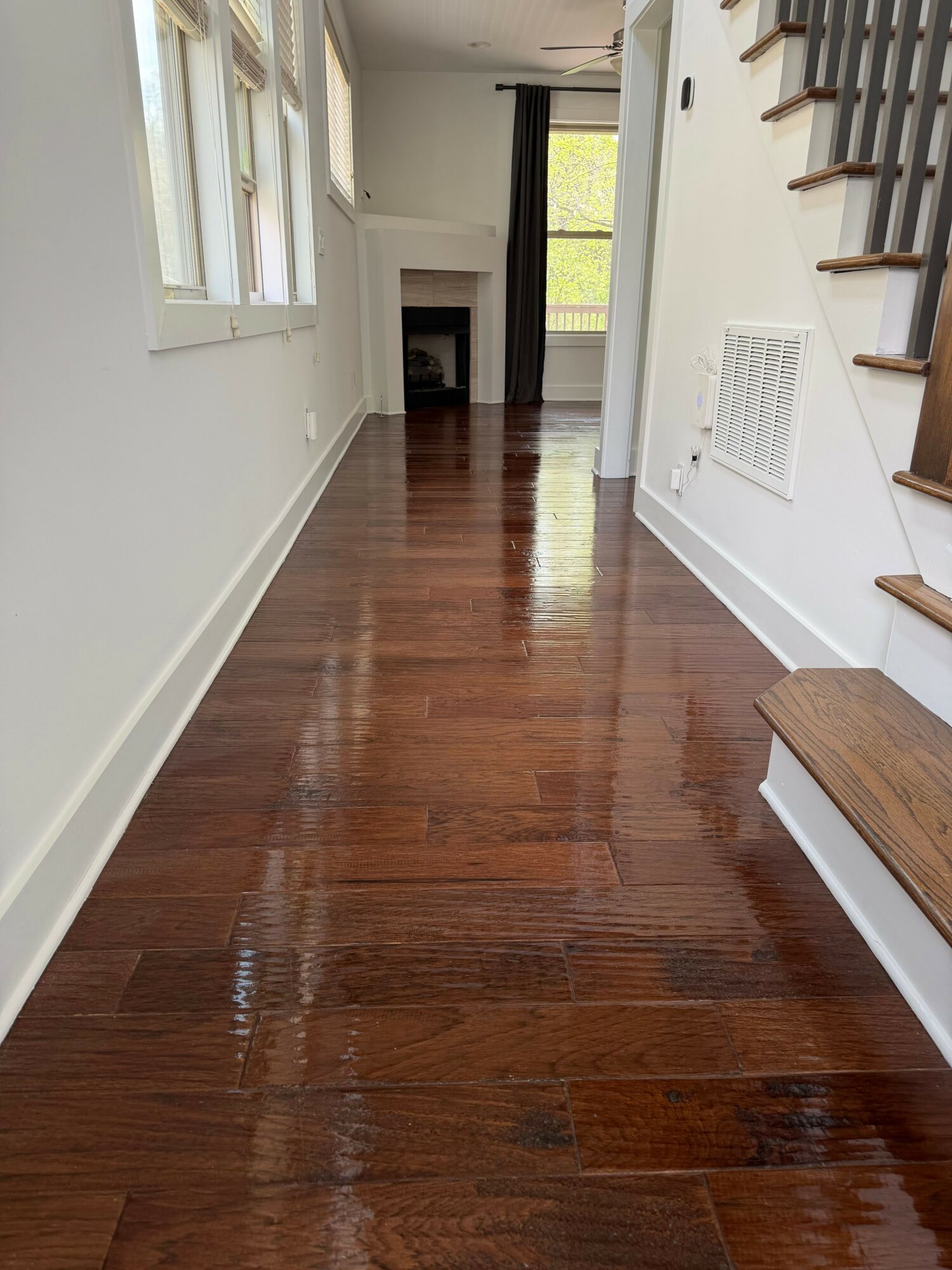 Long hallway with wooden floor, white walls, windows on left, staircase on right, and a fireplace at the end.