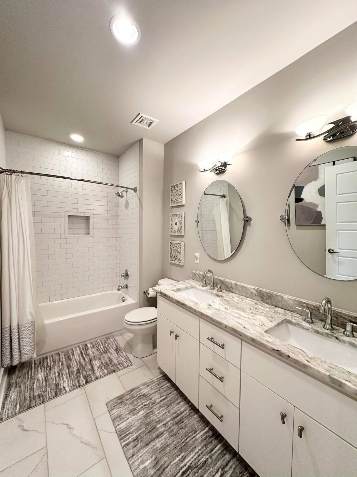Bathroom with bathtub, toilet, double sink vanity, oval mirrors, and framed pictures on wall.