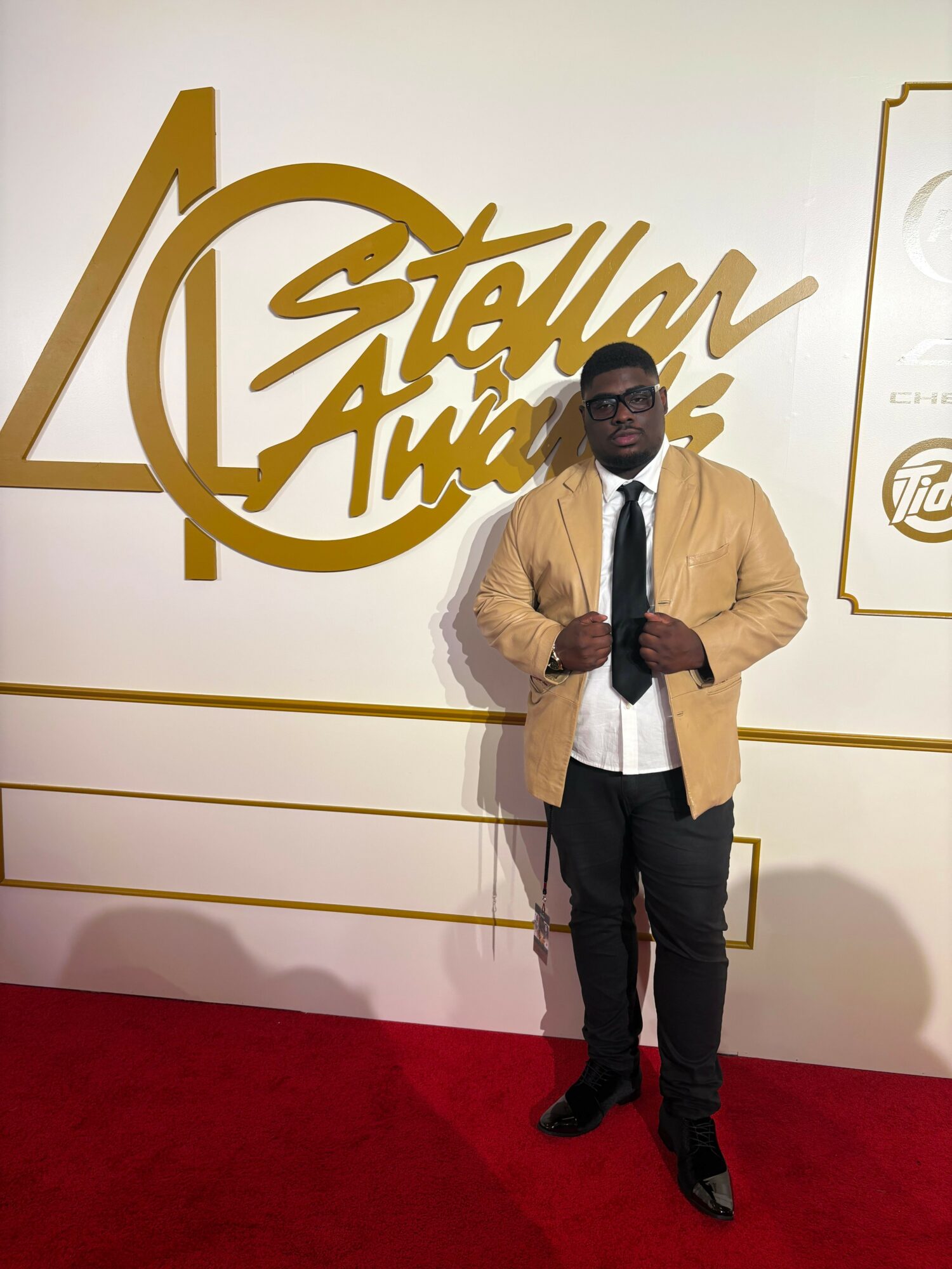 Man in beige jacket, white shirt, black pants, and glasses standing on red carpet in front of a gold logo and white wall.