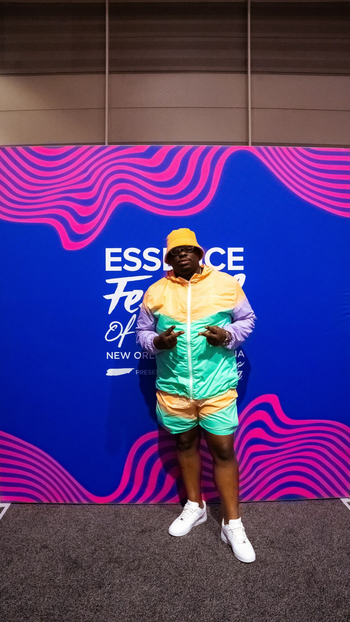 Person standing in front of a blue and pink patterned backdrop, wearing a yellow hat, colorful jacket, shorts, and white shoes.