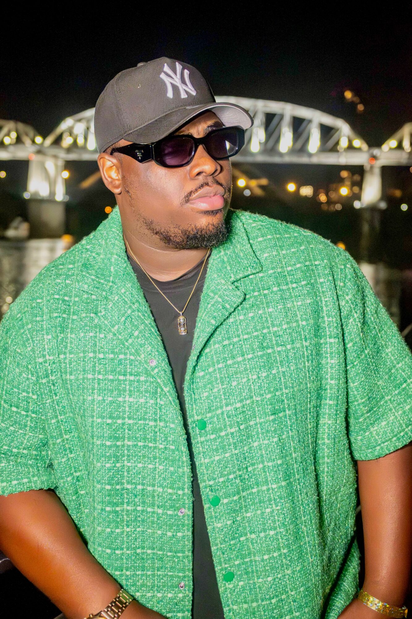 Man wearing sunglasses, baseball cap, green shirt, and jewelry, with a bridge and city lights in background.