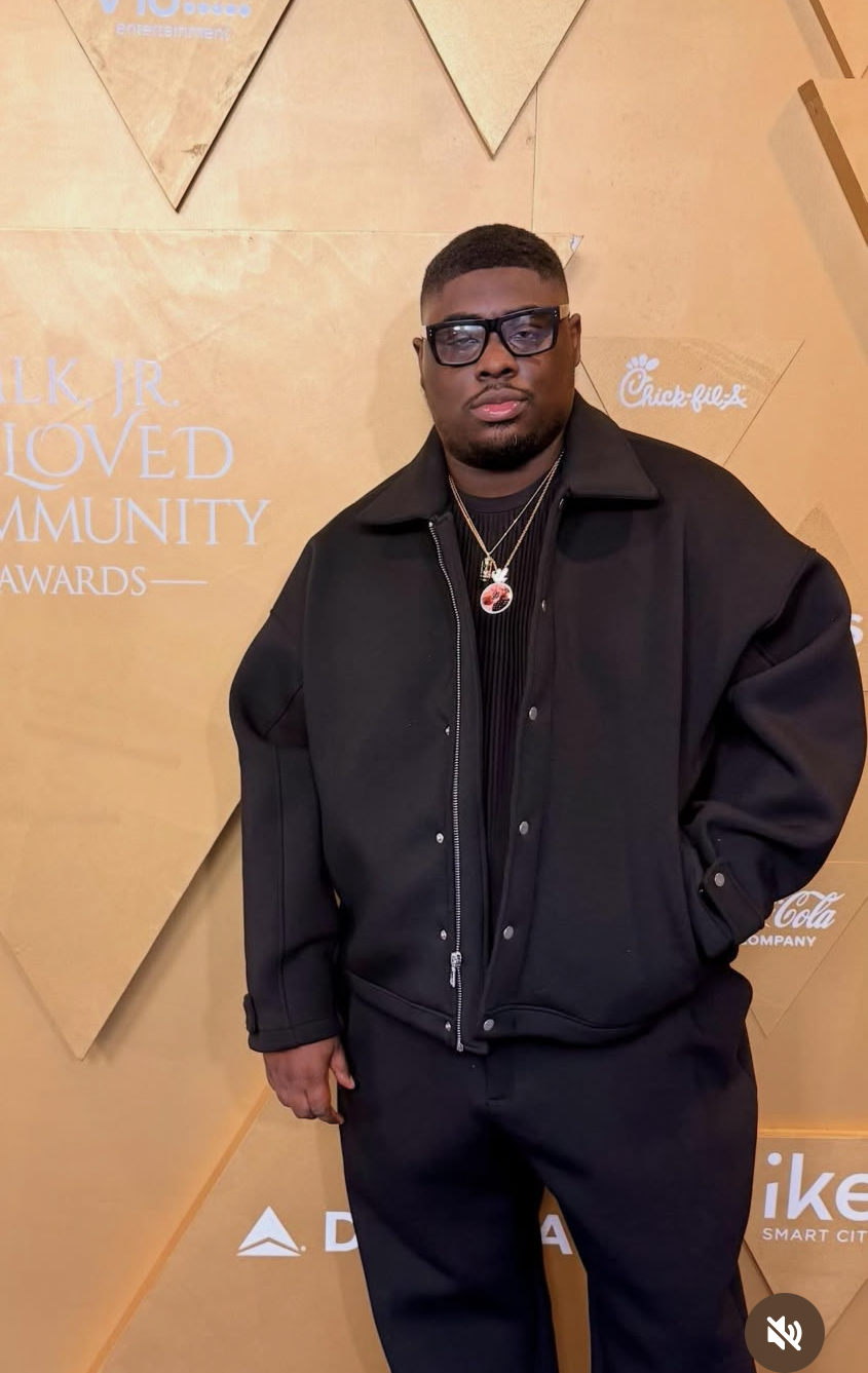 Young man with glasses and jewelry standing in front of a beige backdrop with text and logos.