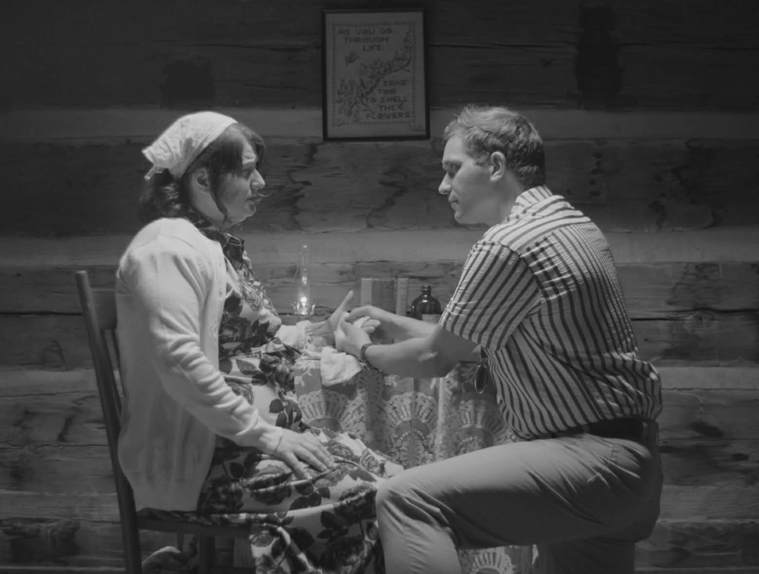 A man and woman sit facing each other, holding hands in a room with wooden walls and a framed picture.