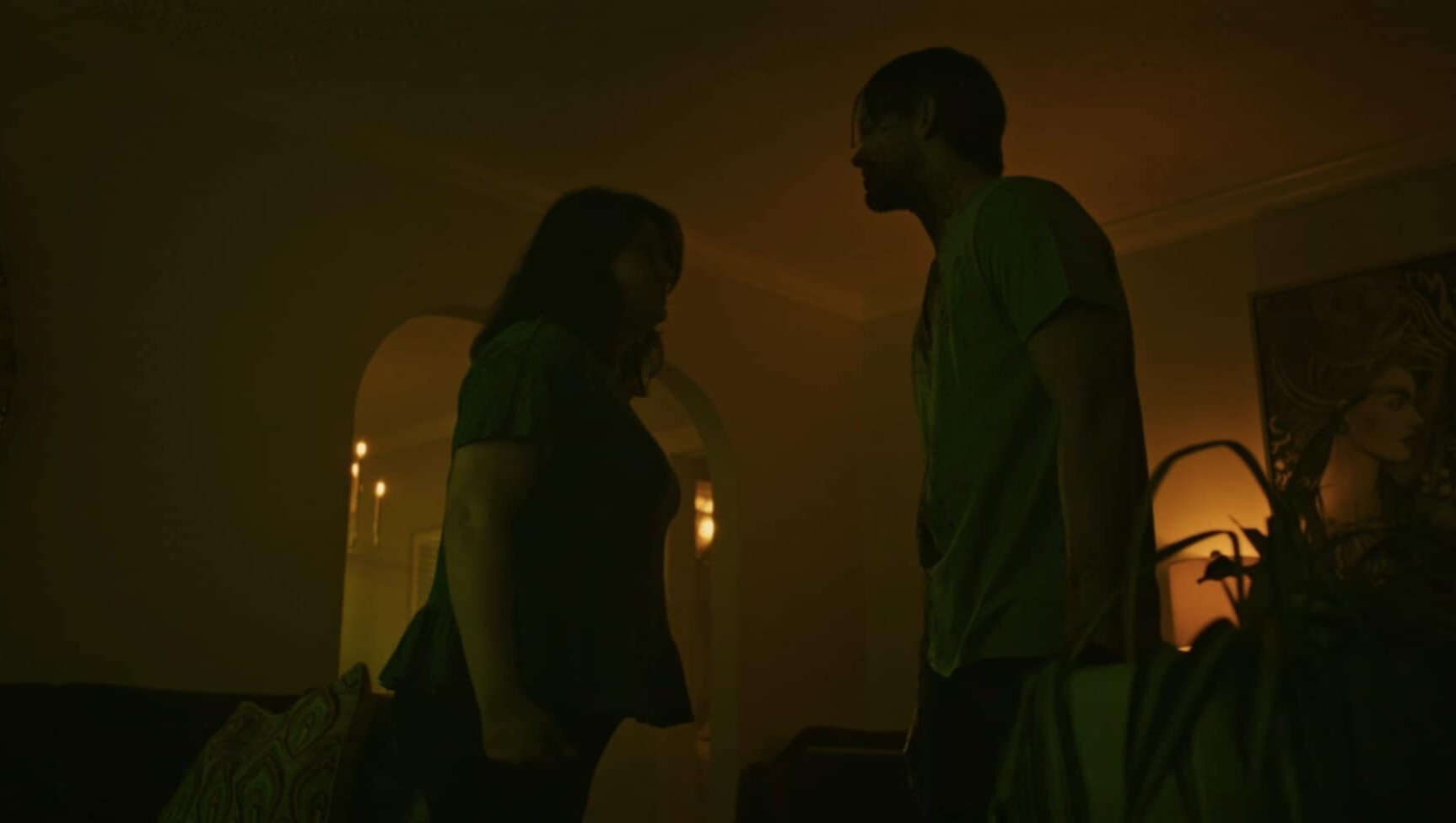 Two people standing indoors, facing each other in a dimly lit room with warm lighting and a plant nearby.