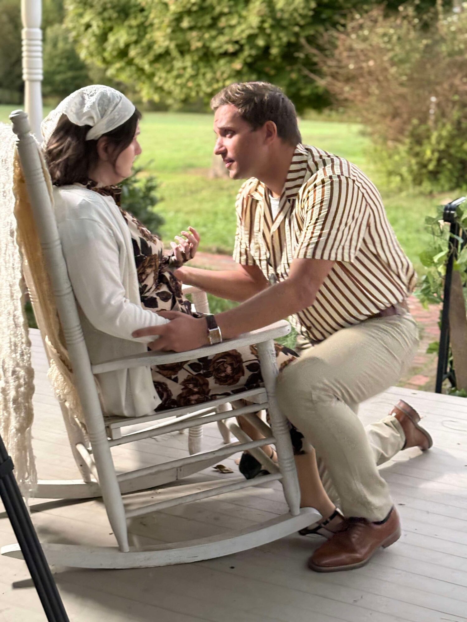 Man kneeling and talking to woman sitting on a porch swing outdoors with greenery in background.