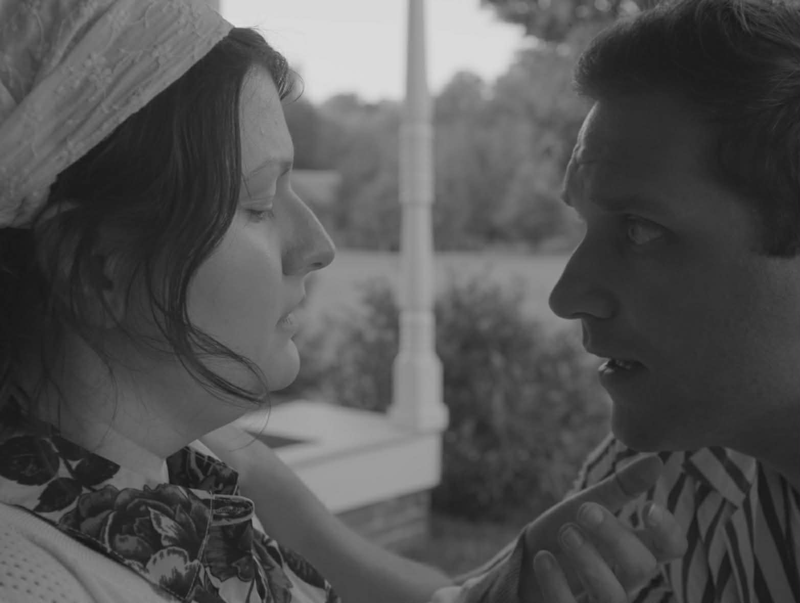 Two people face each other closely outdoors, one with a hat, the other with short hair, engaging in conversation.