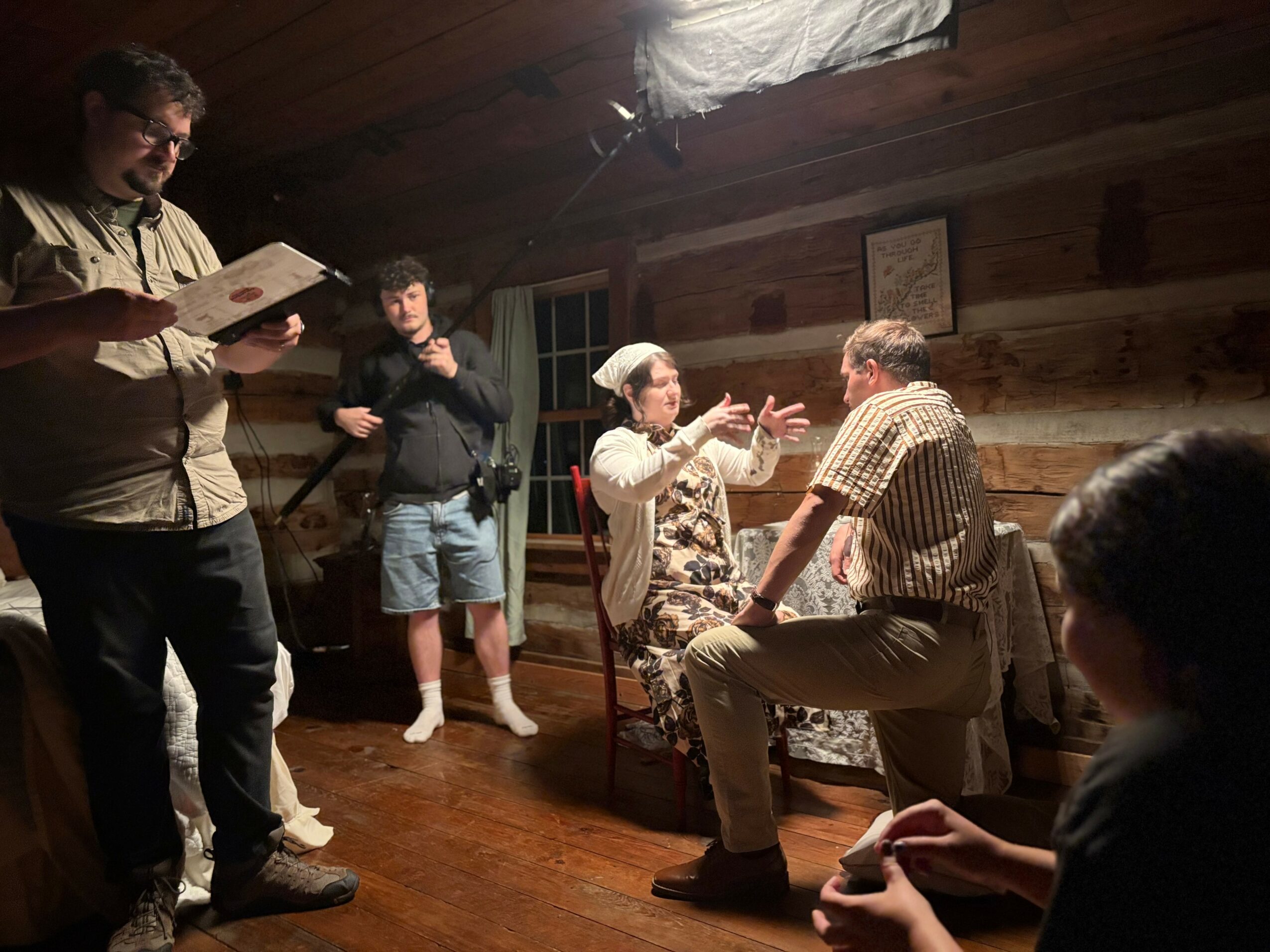 Four people in a wooden room, two seated and two standing, engaged in conversation and reading. One person reads a book.
