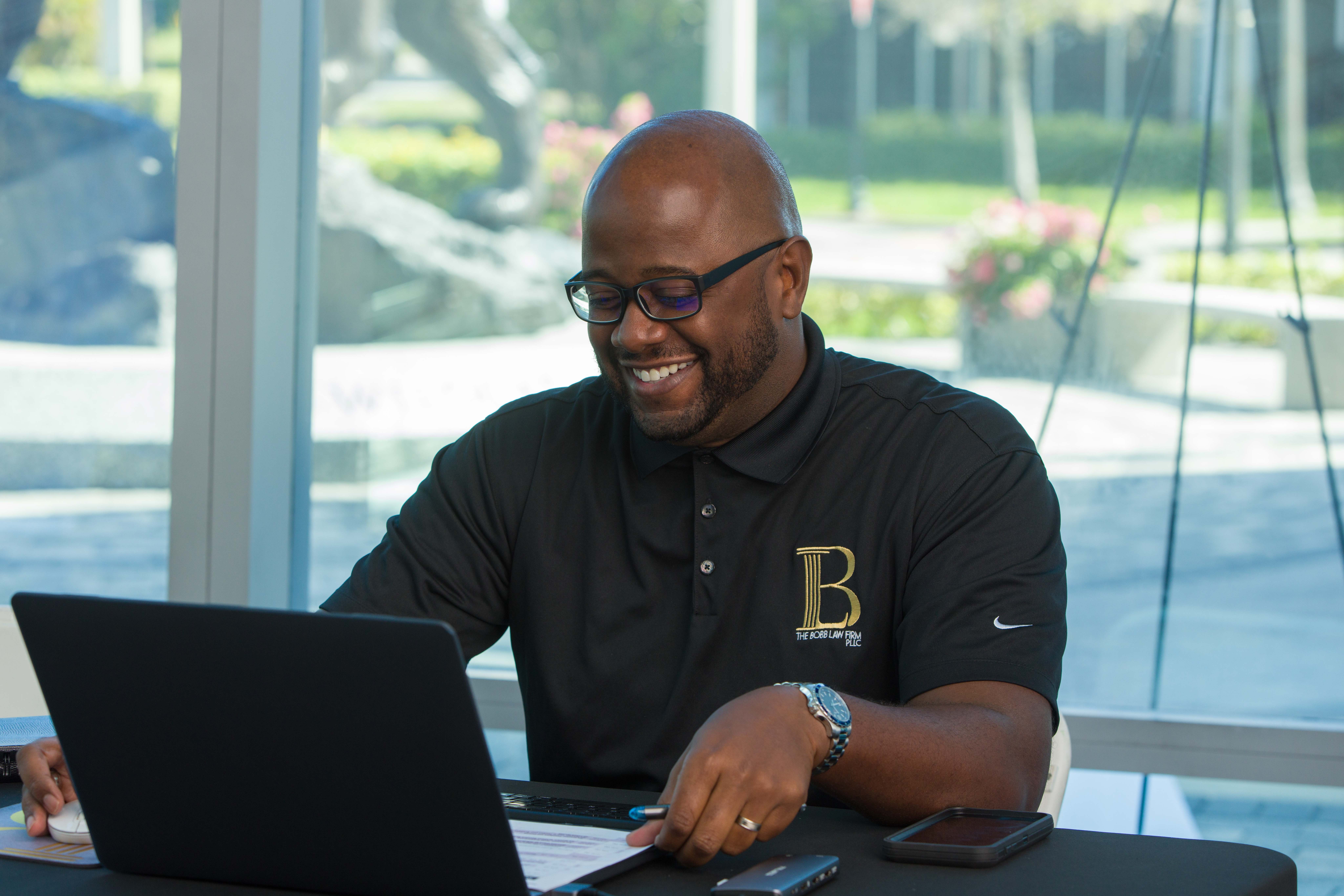 Man with glasses smiling at laptop in bright room with large windows and outdoor scenery.