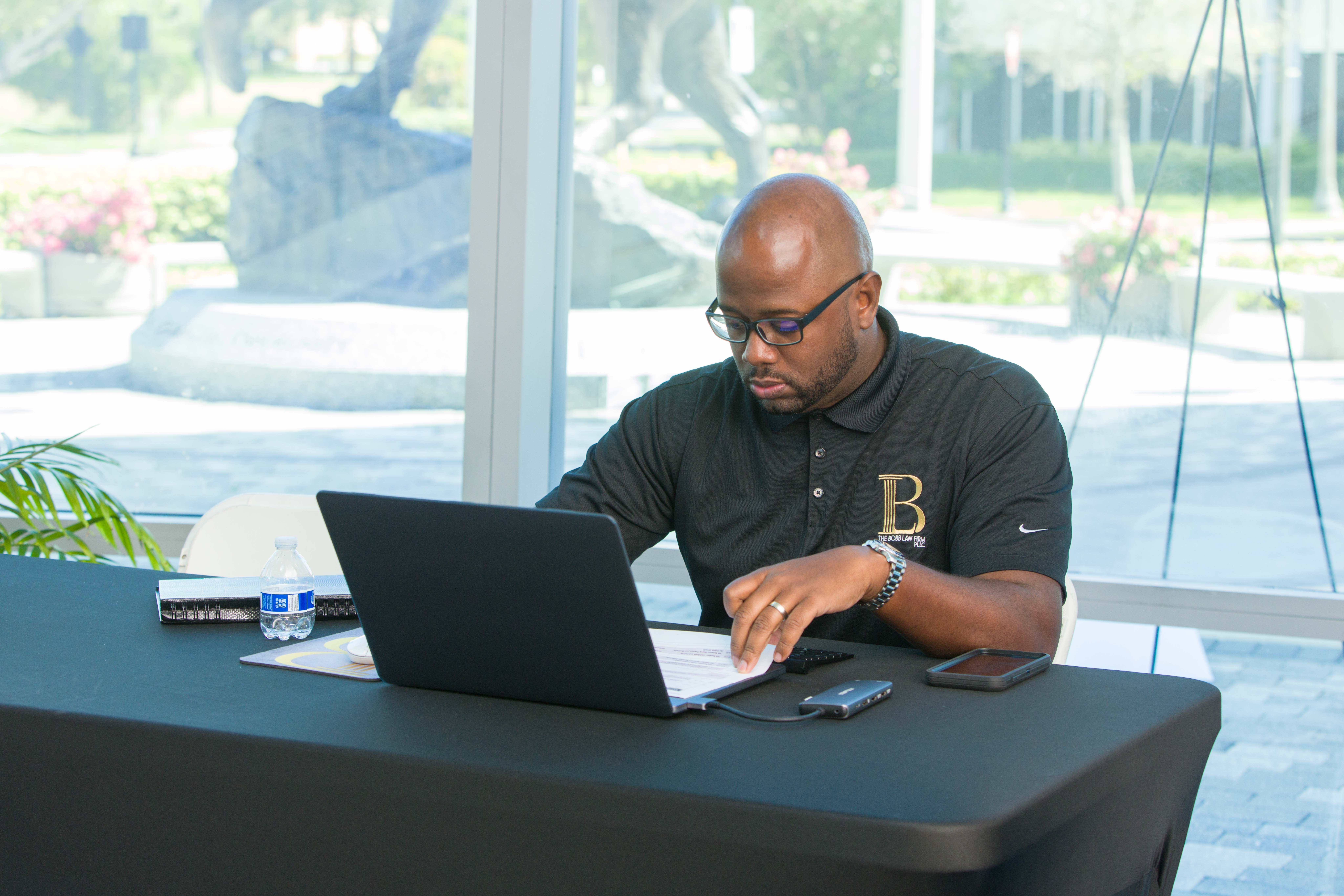Man with glasses working on a laptop at a table in front of large windows, with a mobile phone nearby.