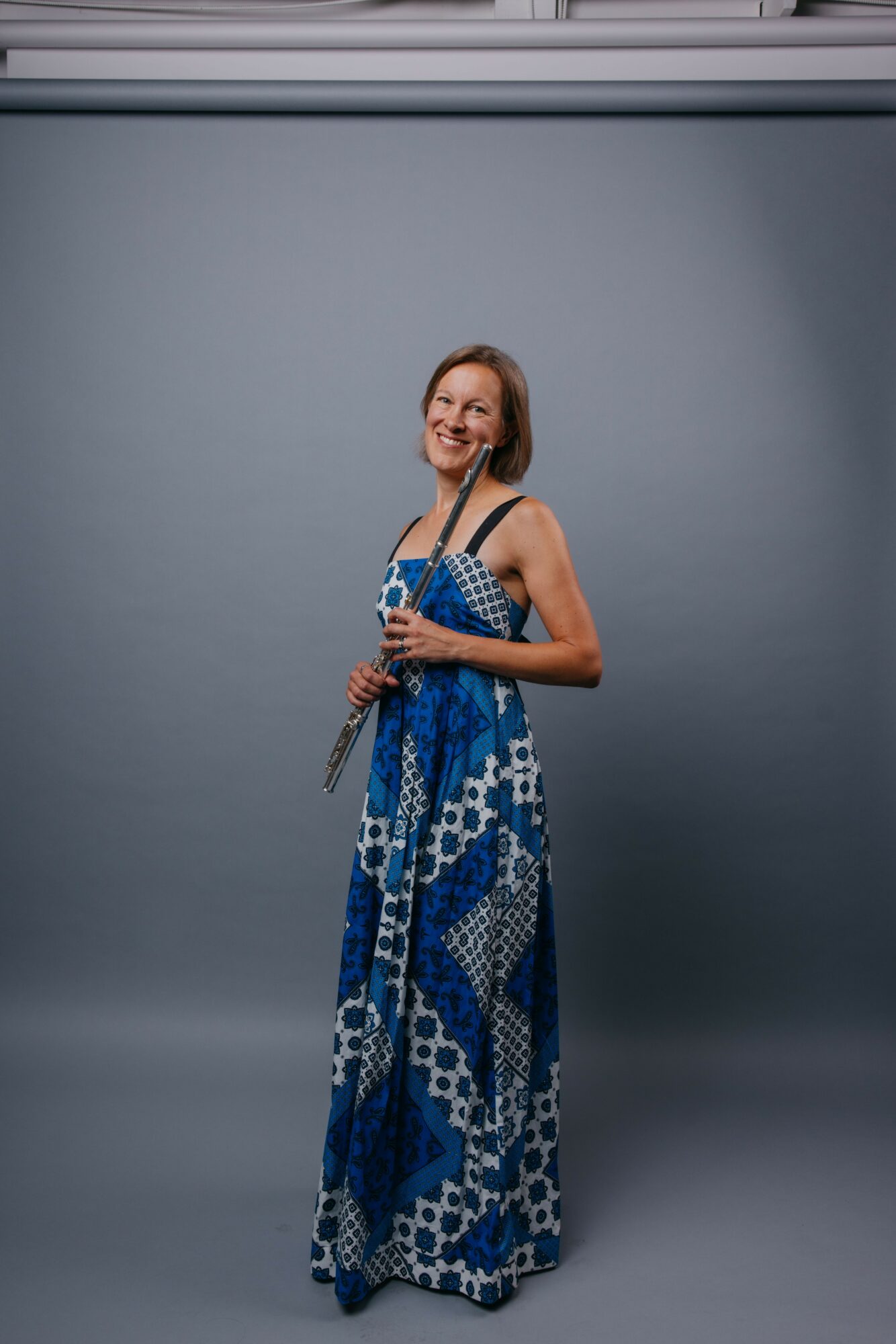 Woman in a blue patterned dress holding a camera, smiling, standing against a plain gray background.