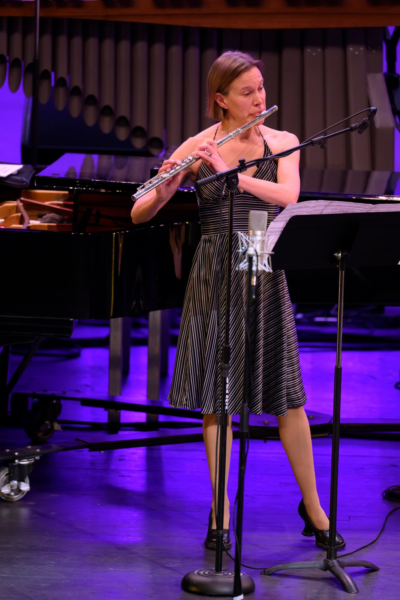 Woman playing flute on stage with piano and music stand, purple lighting, wooden backdrop.
