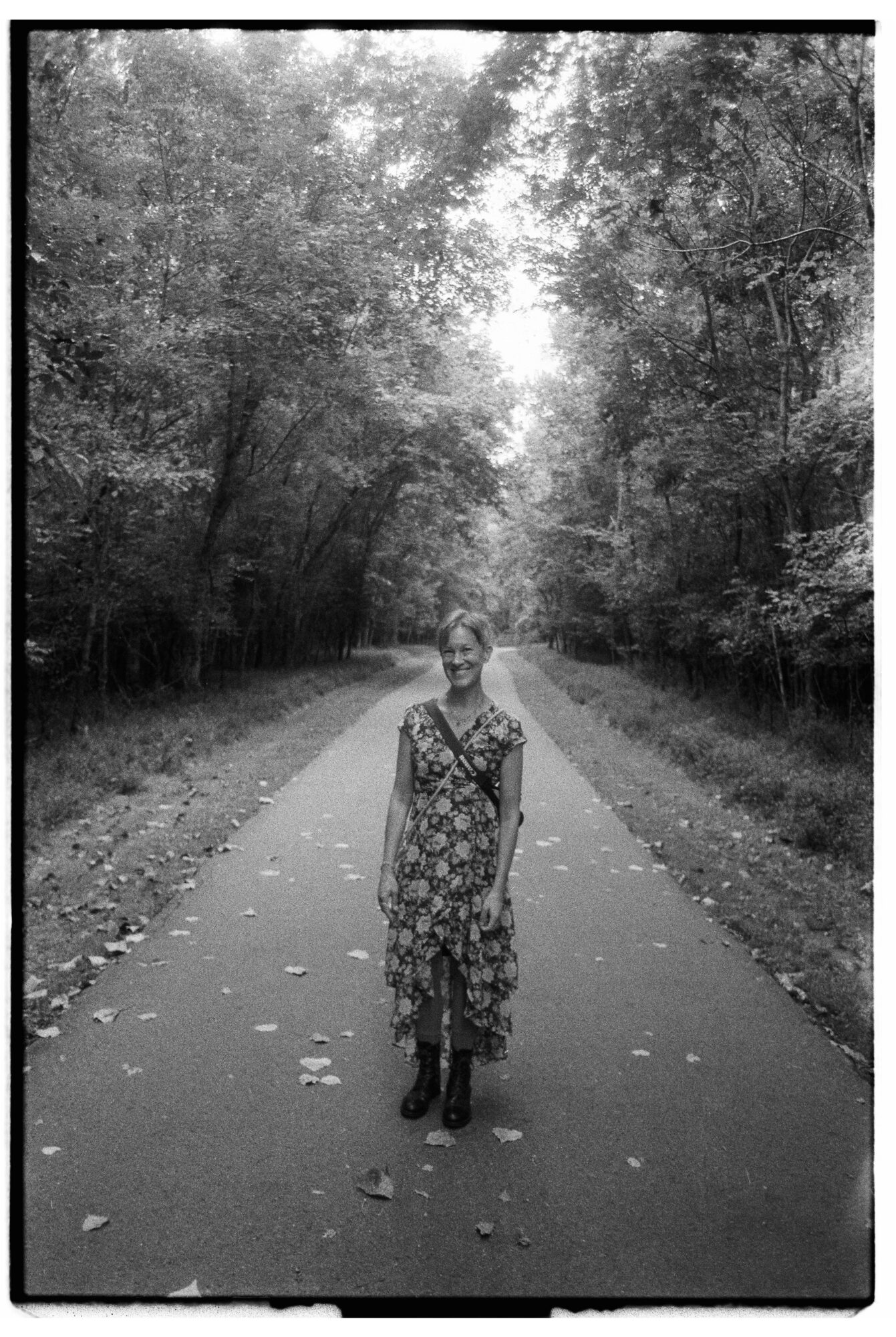 Person standing on a paved path surrounded by trees, with leaves scattered on the ground, in a forest setting.