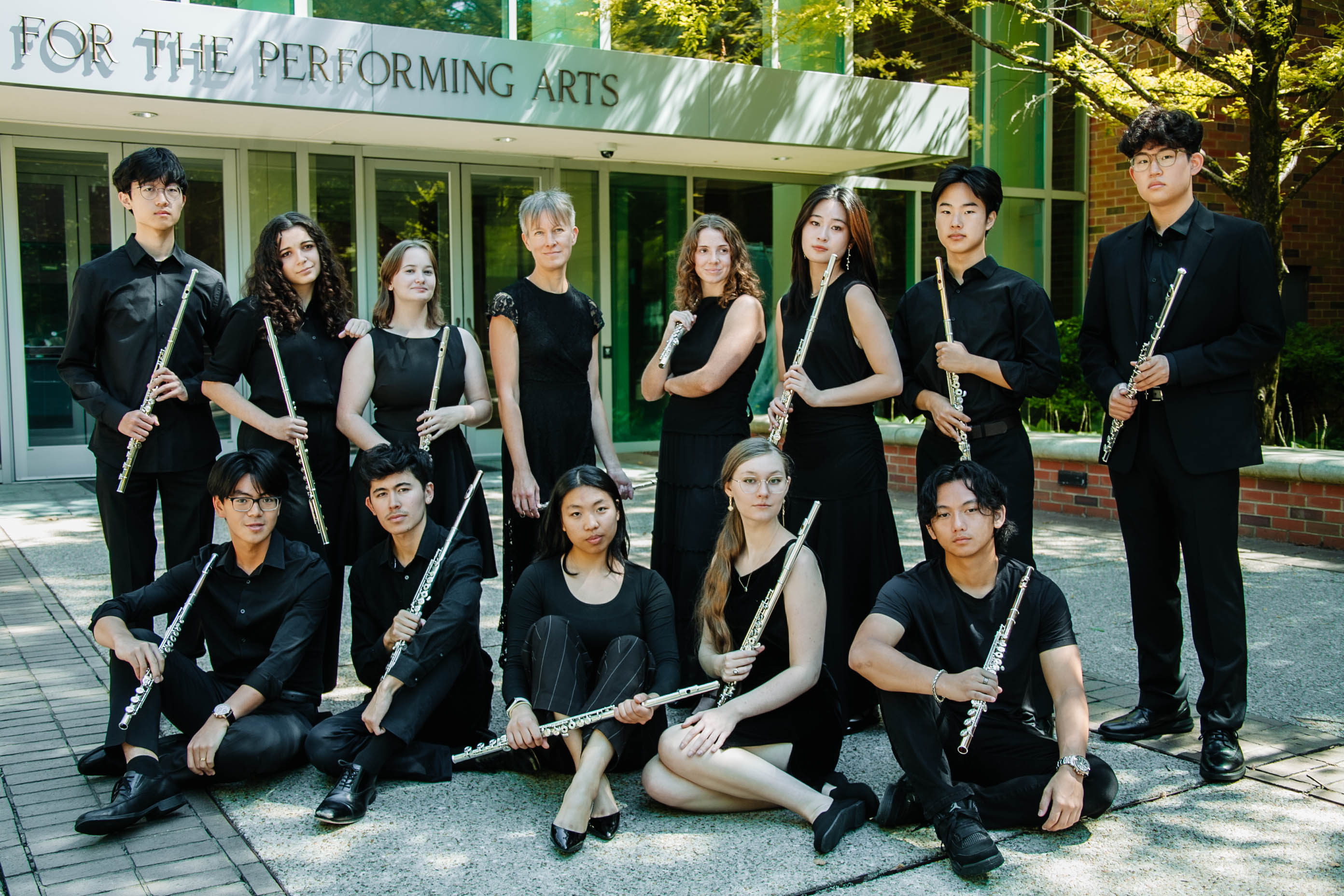 Group of twelve young musicians holding flutes outdoors in front of a building with a sign for performing arts.