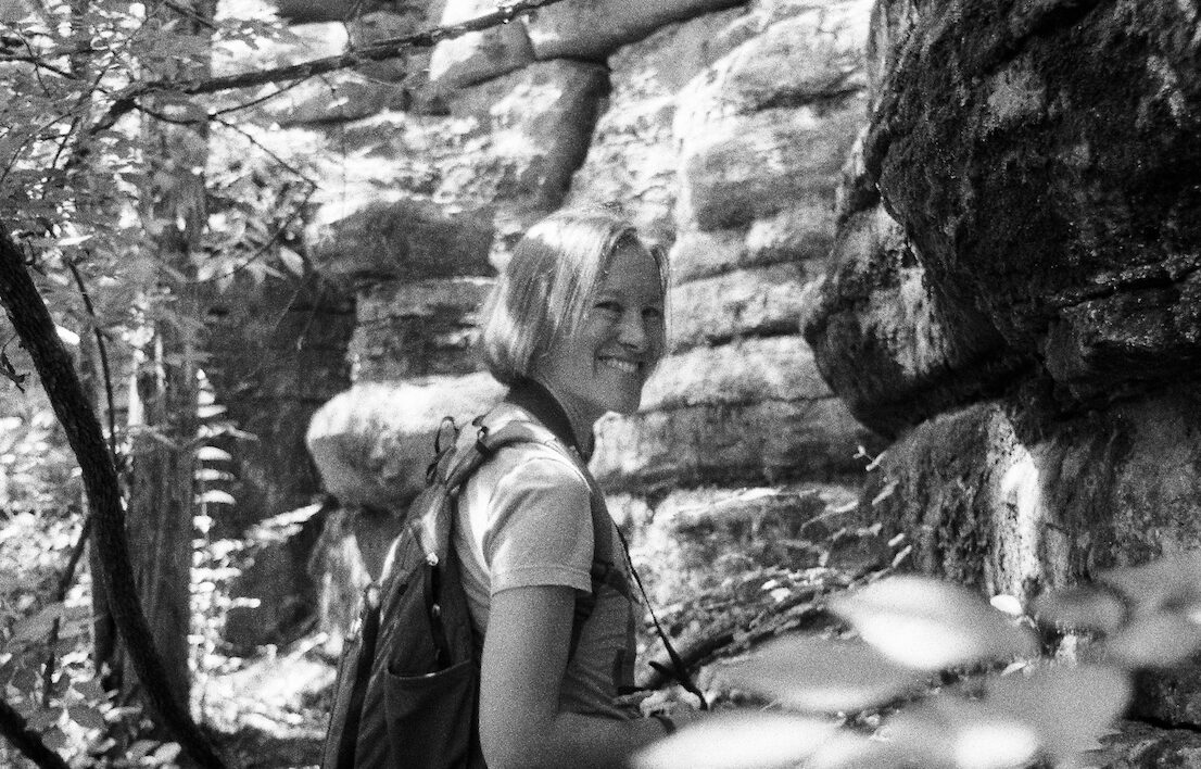 Person with short hair and backpack hiking near rocky cliff in forest, smiling.