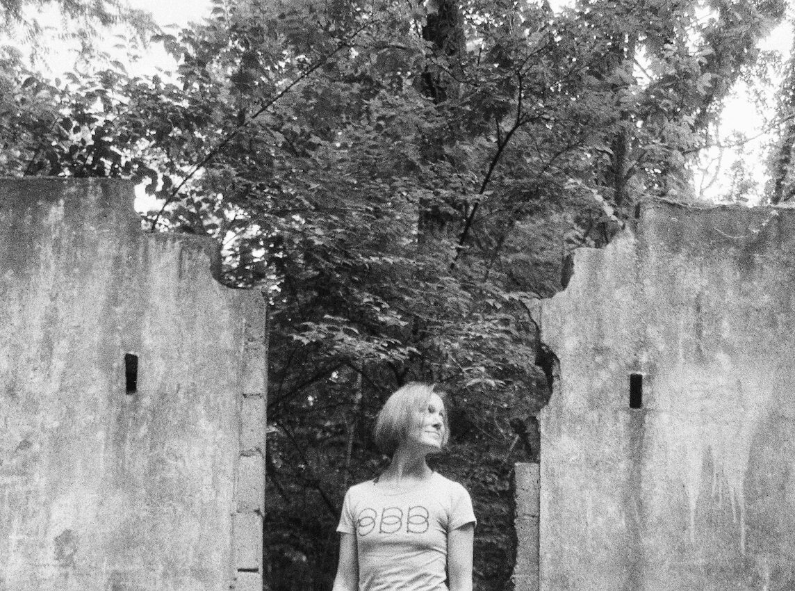 Young girl with short hair standing in front of a large tree and concrete wall.