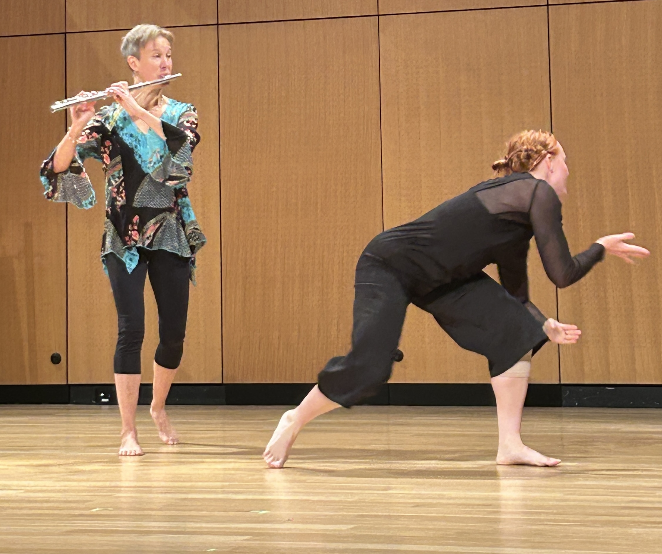 Two women perform on stage with a wooden panel background; one playing flute, the other in a crouched pose.