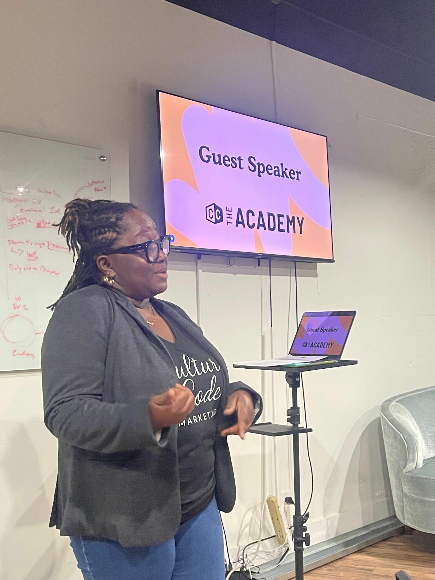 Woman speaking in front of a screen displaying 'Guest Speaker' and 'Academy' at an indoor event.