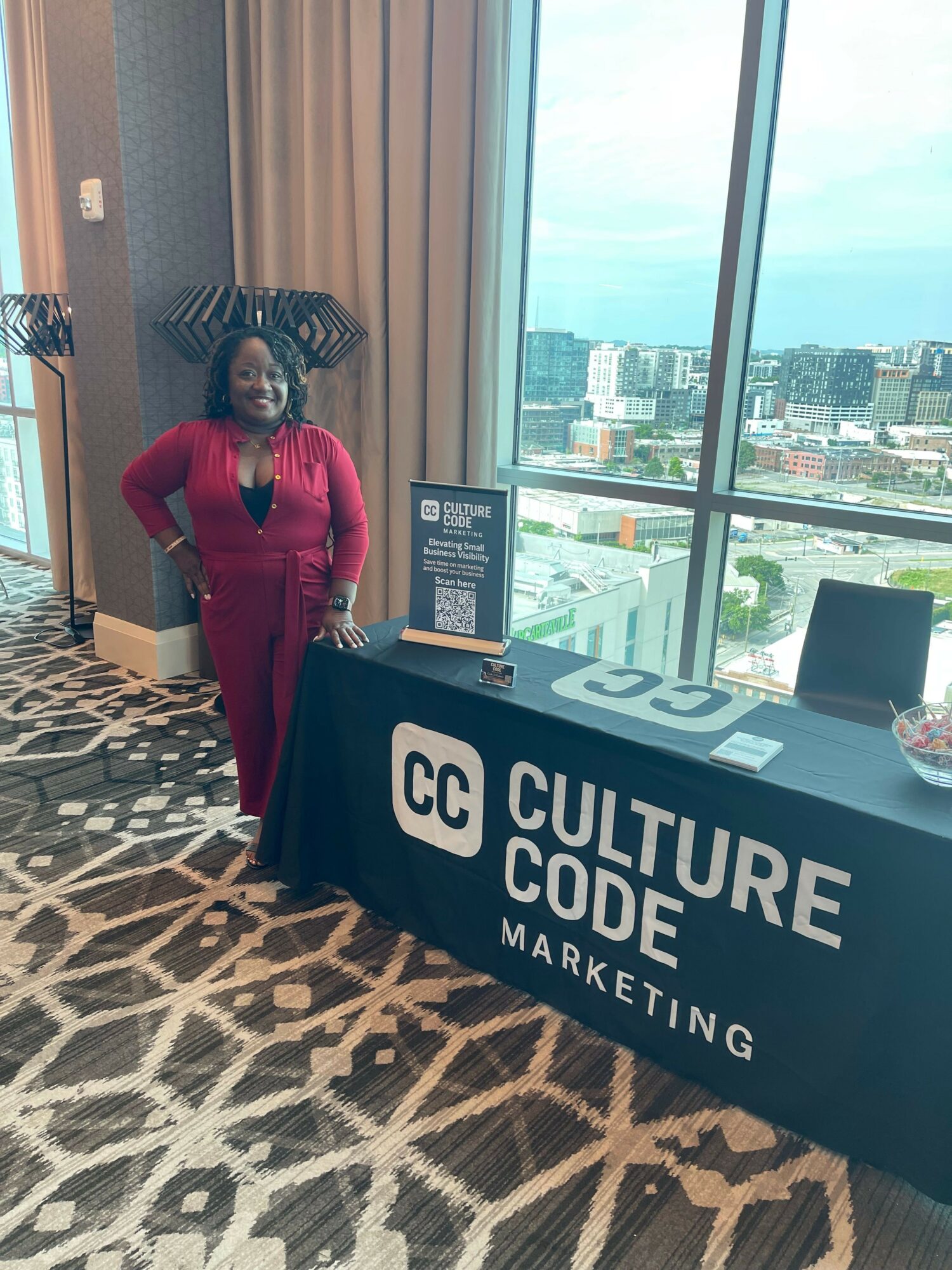 Woman in red standing next to a table with a sign and promotional materials, high-rise city view through window.
