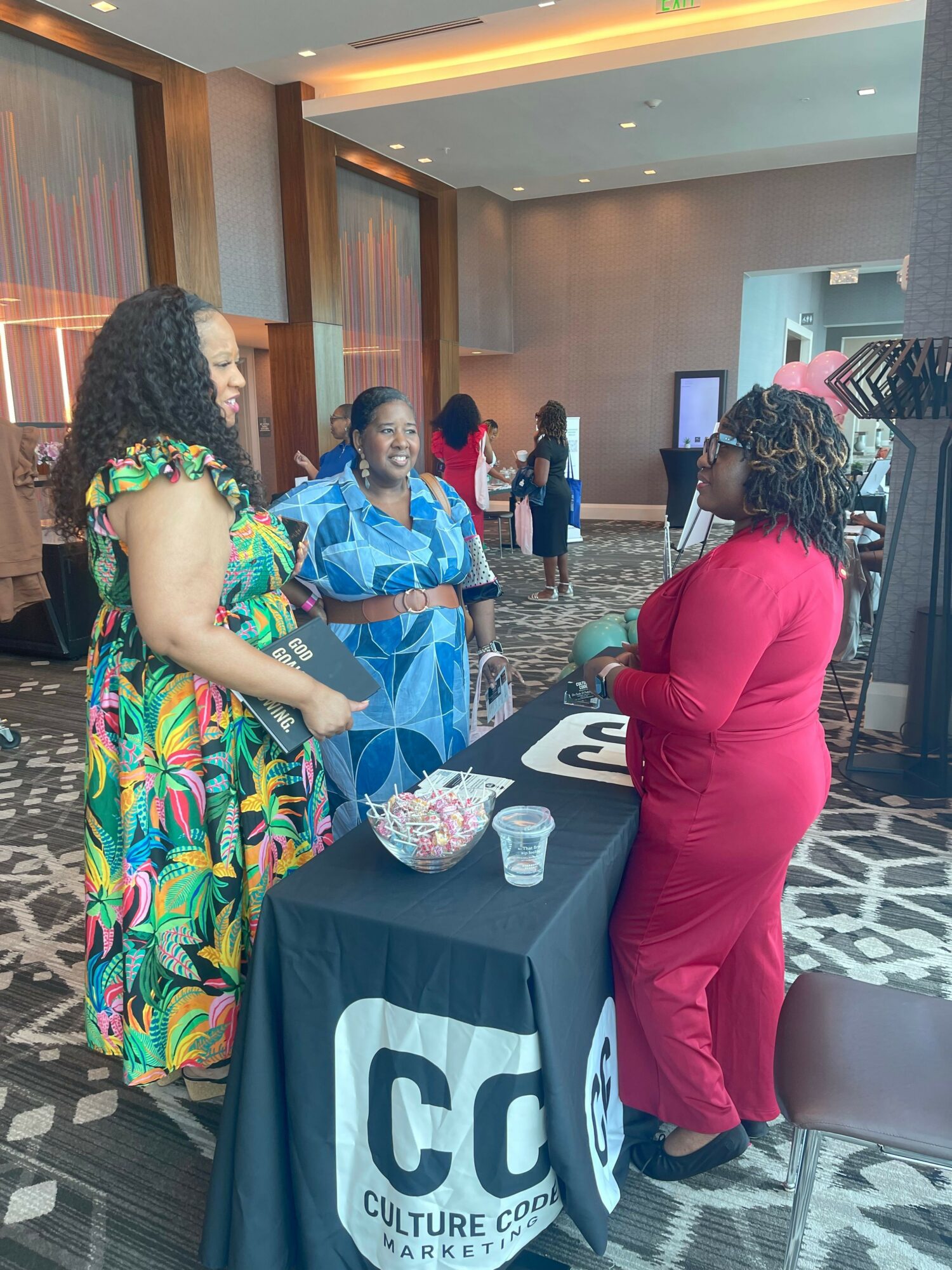 Three women engaged in conversation at a table with a bowl of snacks and drinks in a conference room.