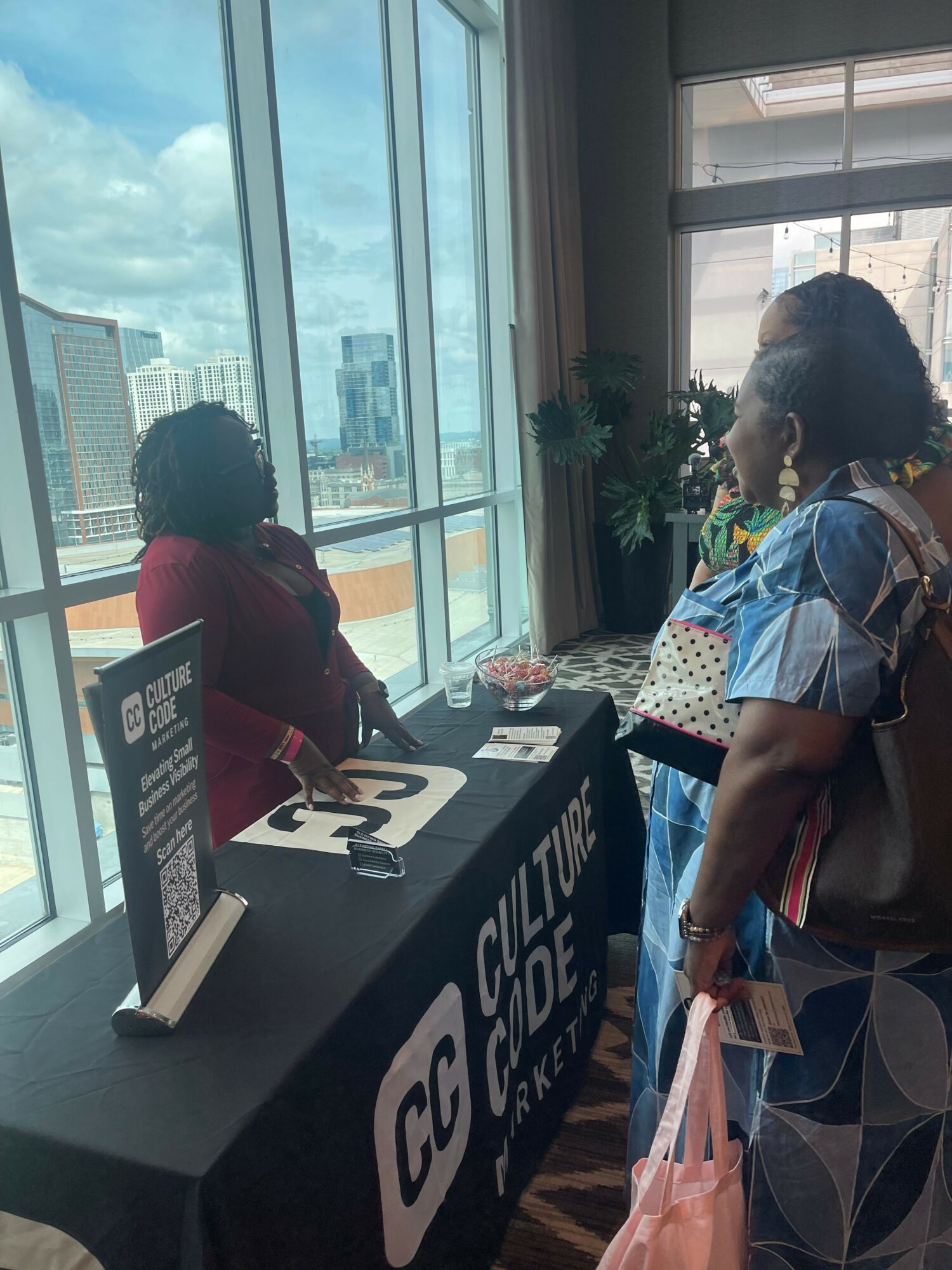 Two women talking near a table with informational materials inside a building with large windows and city view.
