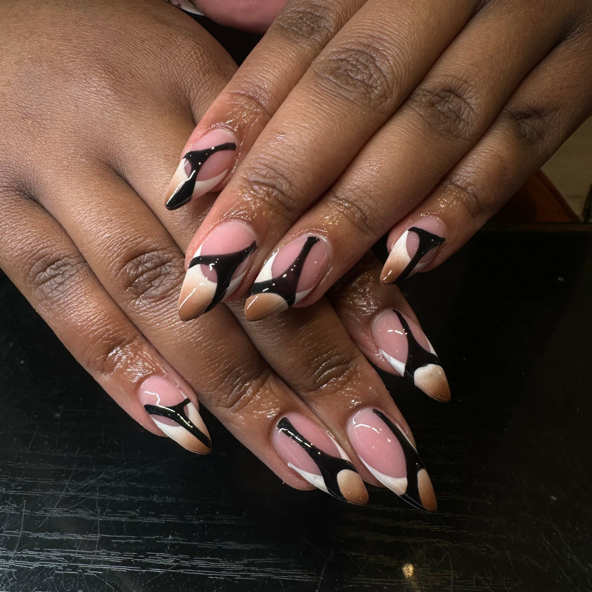 Hands with manicured nails featuring black, pink, and nude geometric designs, resting on a dark surface.
