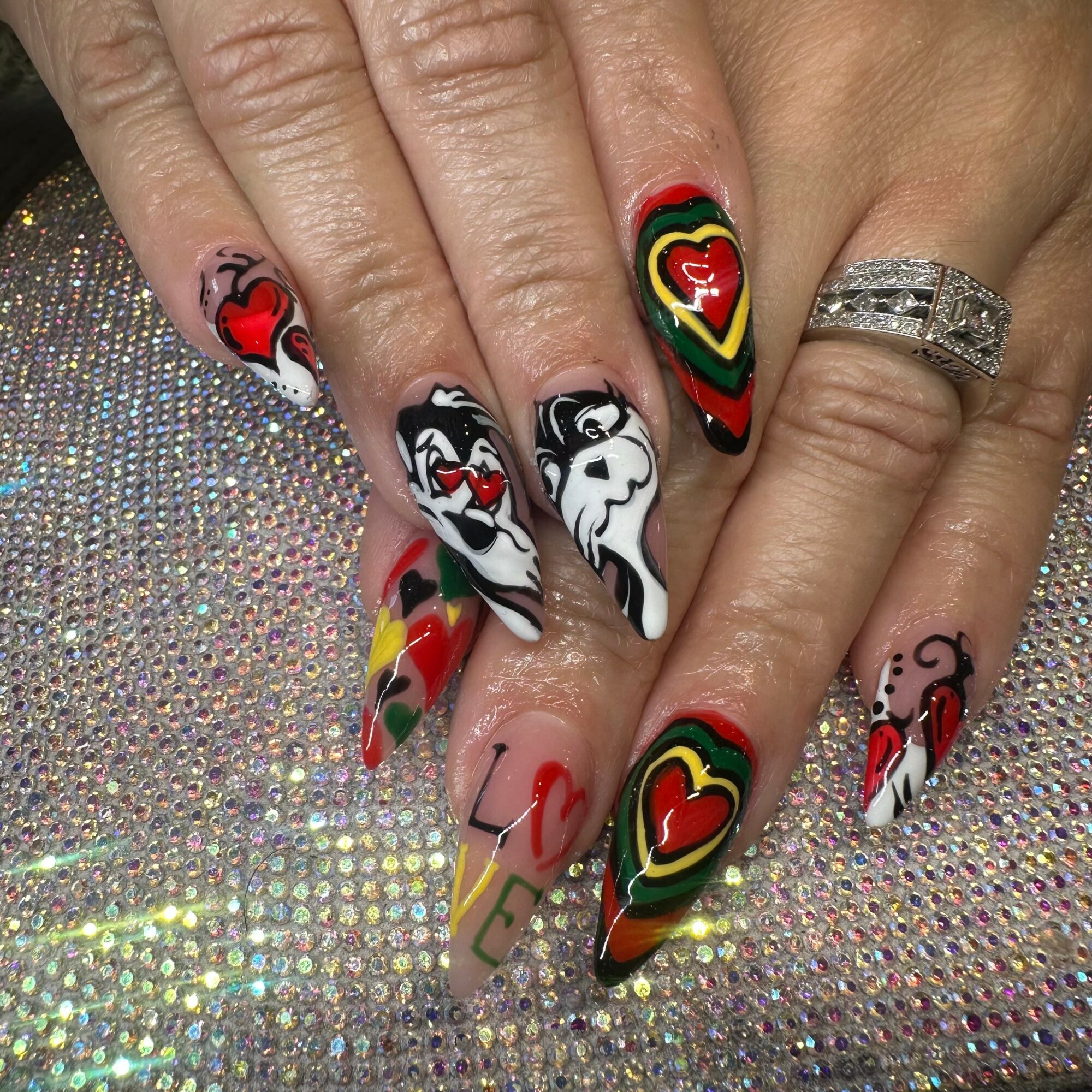 Nail art with colorful hearts, black and white designs, and rainbow stripes on long nails, with a ring on the finger.