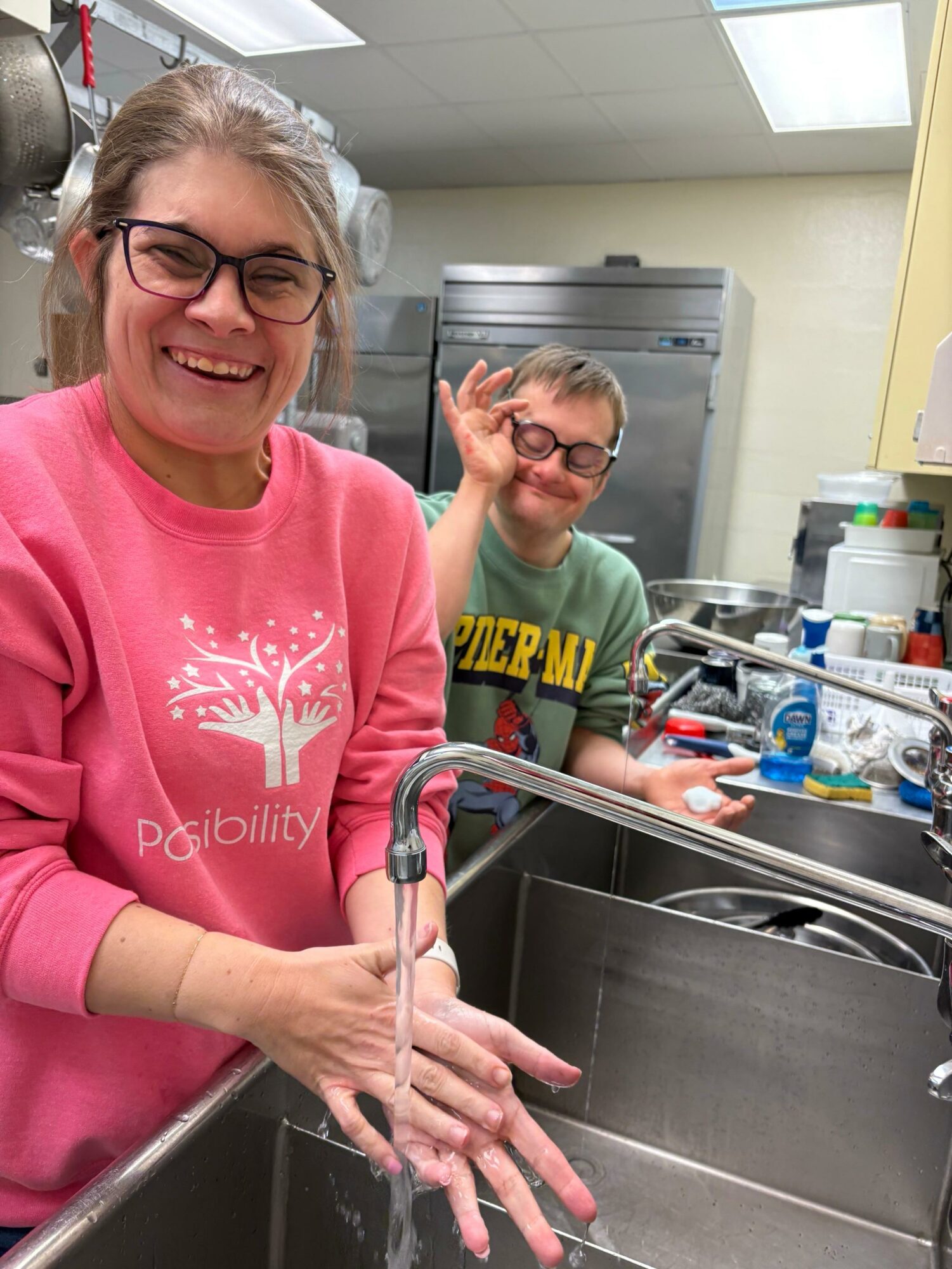 Two smiling people in a kitchen, one washing hands and the other making a peace sign.