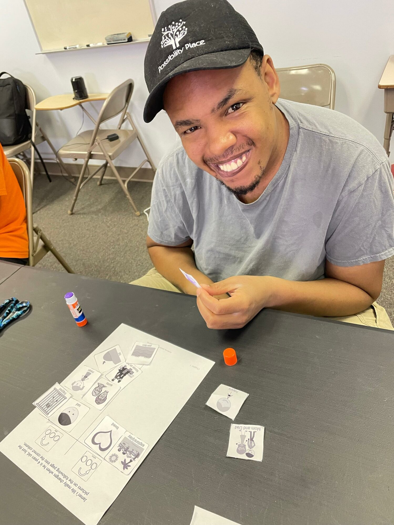 Smiling man wearing a cap and gray t-shirt sitting at a table with game pieces and a paper game board.