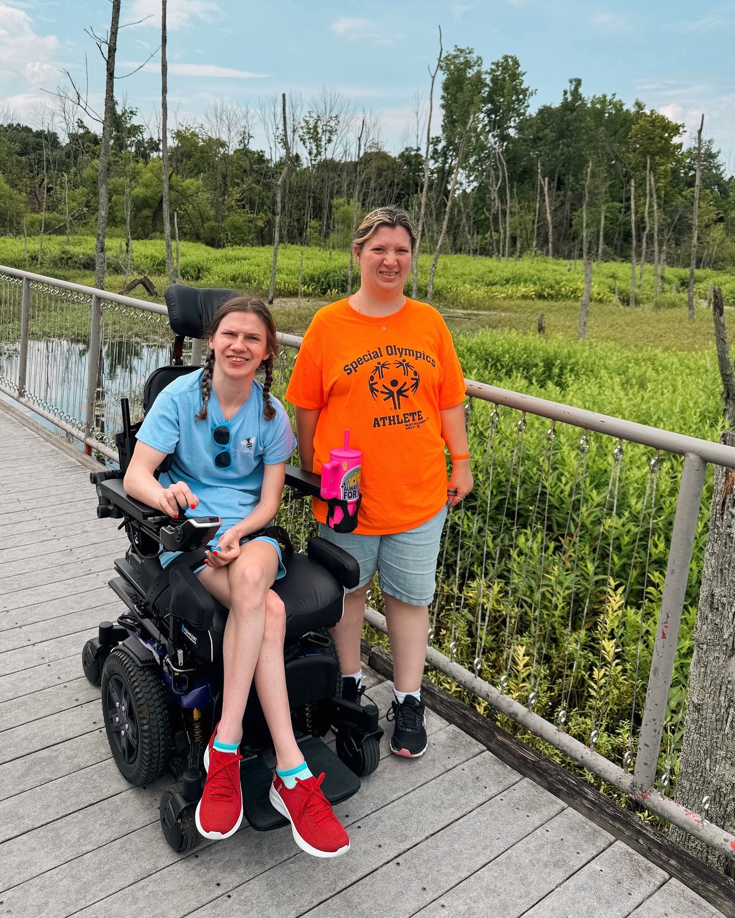 Two people outdoors on a wooden walkway with greenery and trees in the background, one in a wheelchair and one standing.
