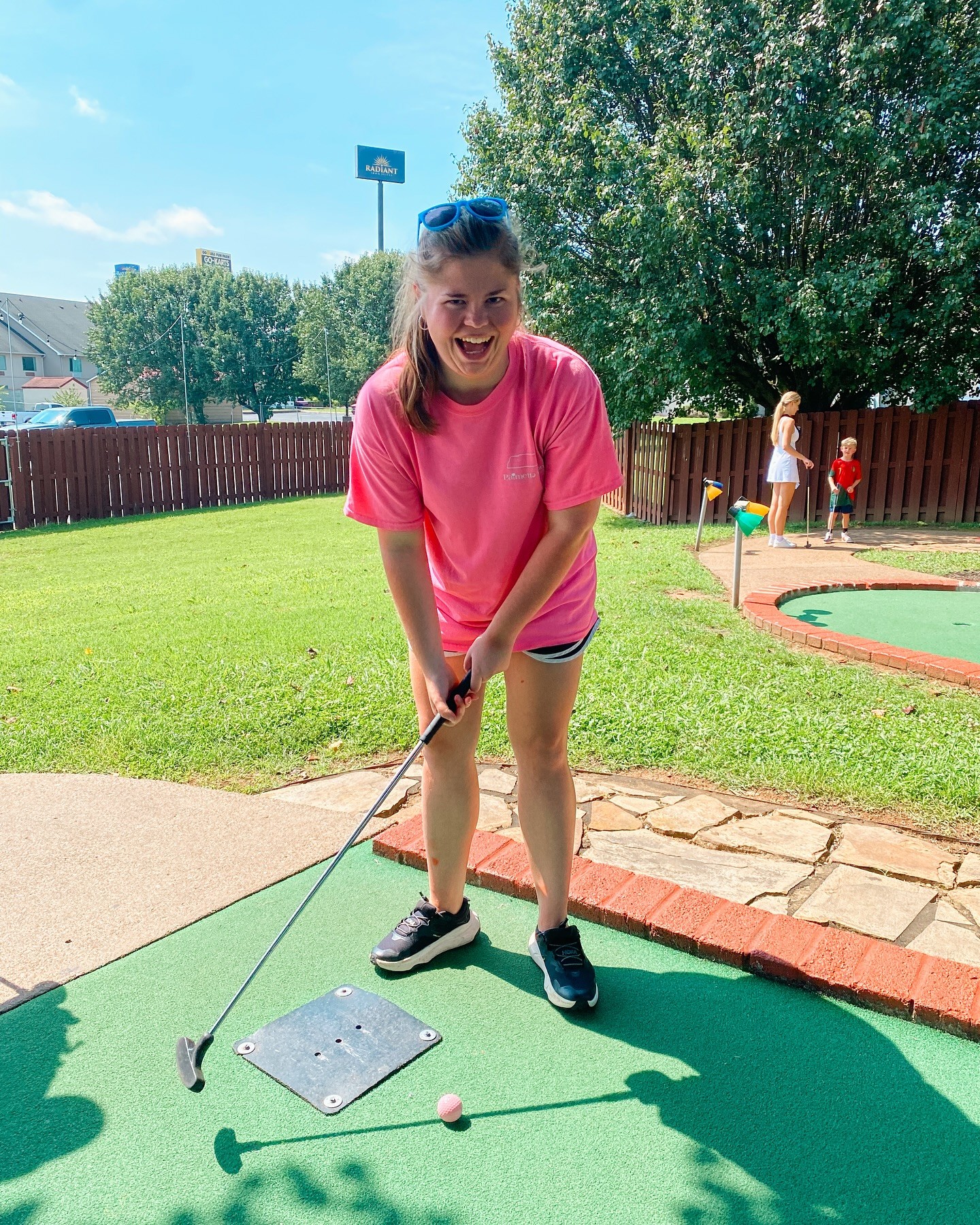 Girl in pink shirt and shorts playing mini-golf outdoors, smiling, holding a golf club, on a green mat with a pink ball.