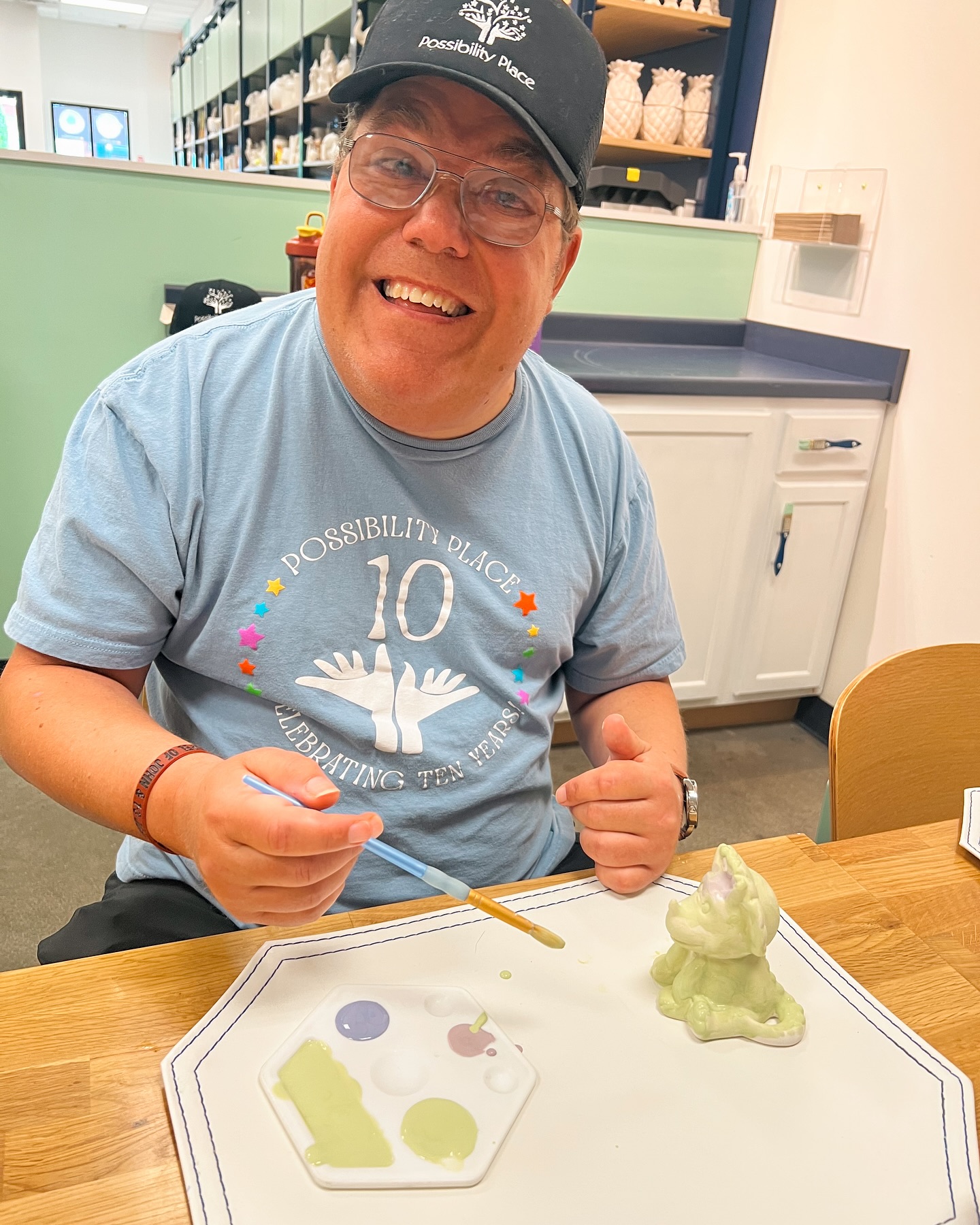 Person smiling, wearing glasses and a cap, sitting at a table with a colorful plate and a green sculpture.