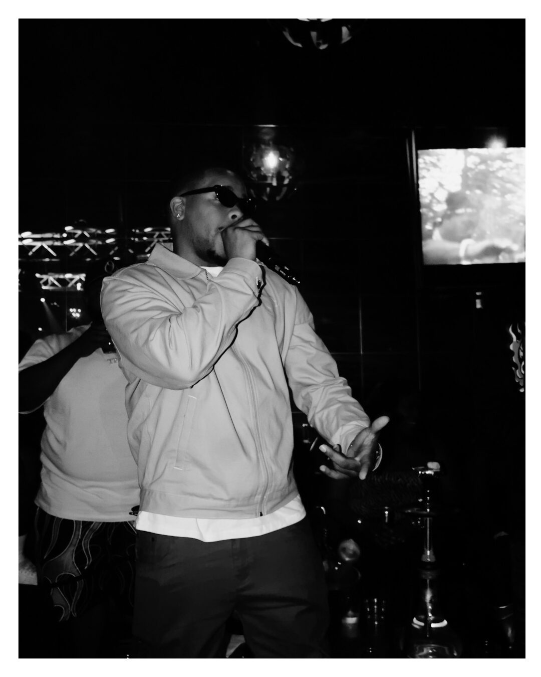 Person singing into a microphone in a dimly lit setting, holding a drink, wearing sunglasses and a jacket.